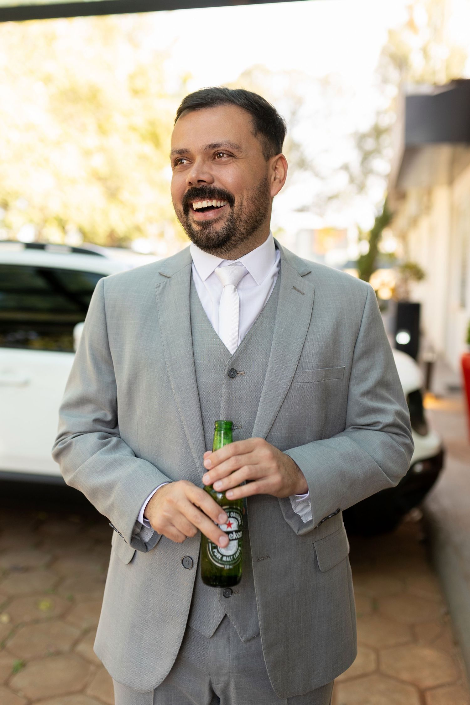 Smiling bearded man in gray three-piece suit holding green beer bottle outdoors.