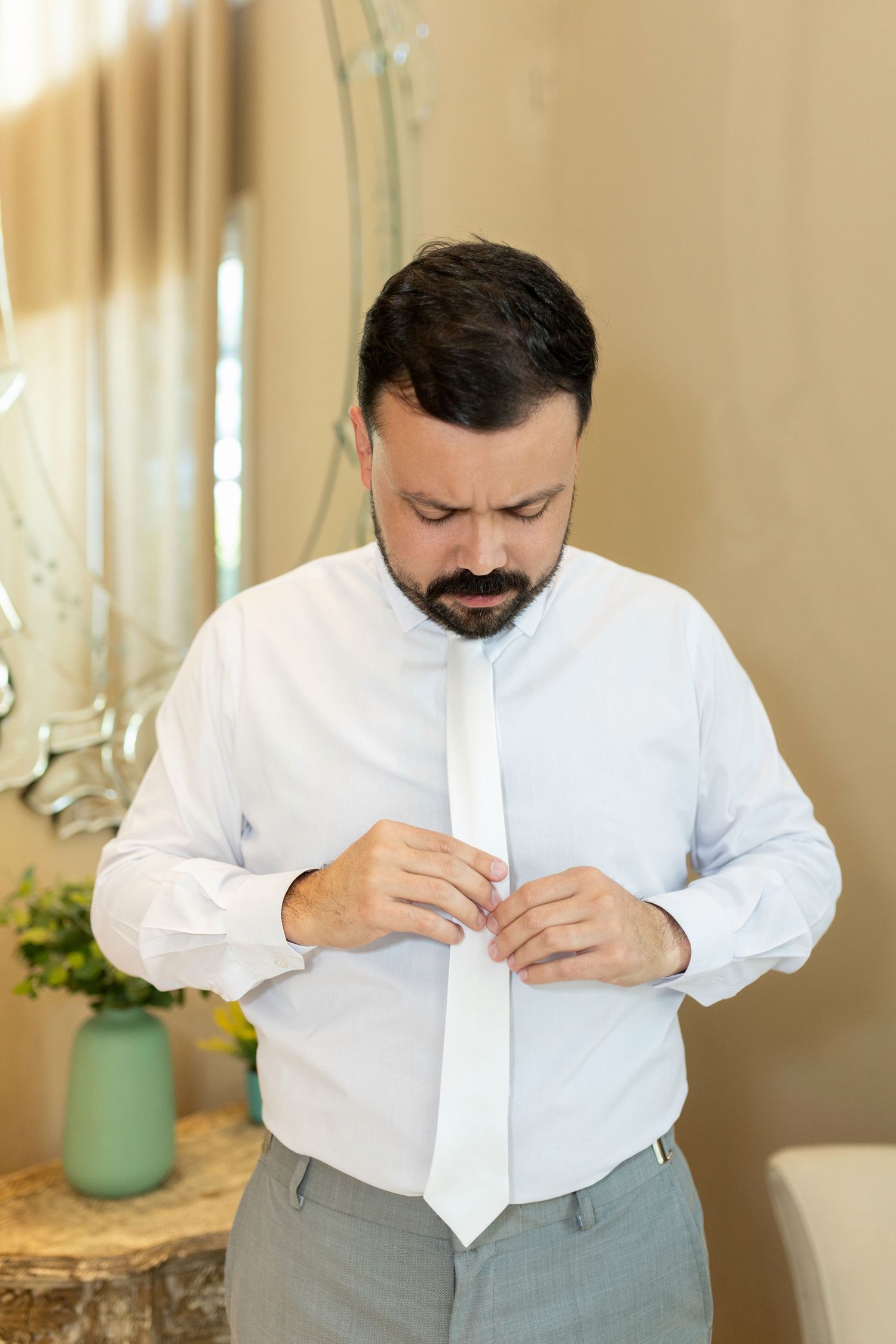 Bearded groom in white dress shirt adjusting his white tie while getting ready for wedding ceremony.