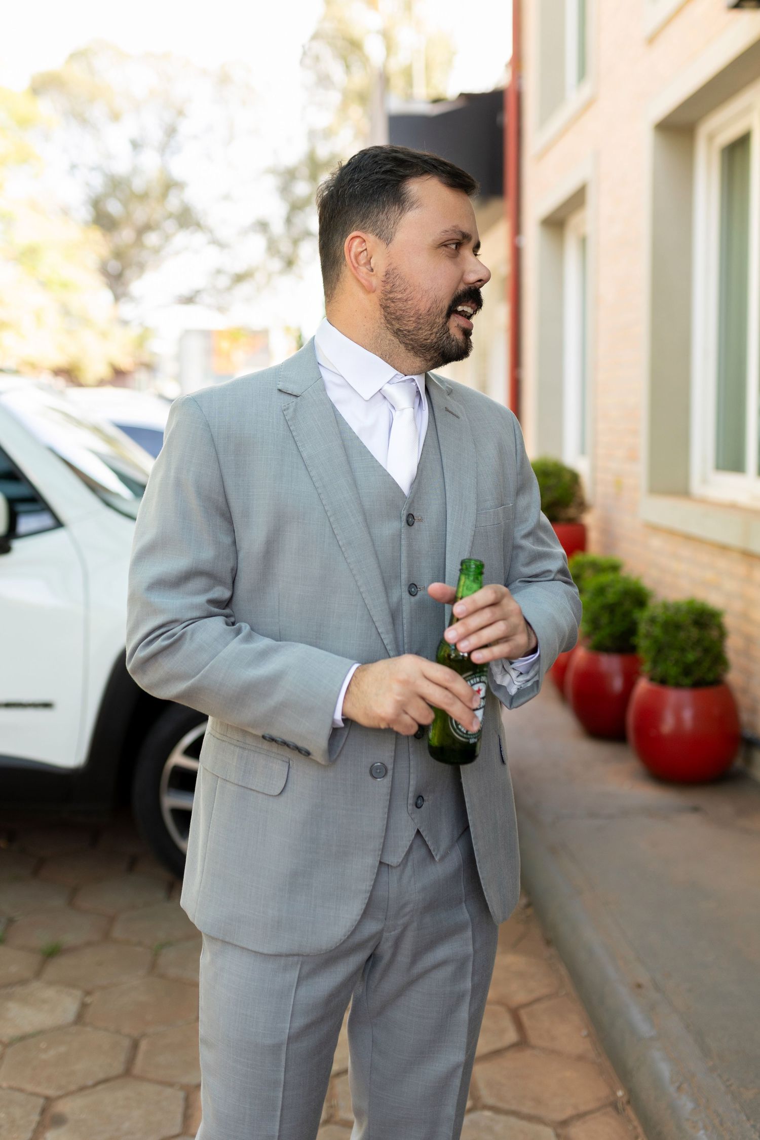 Man in gray three-piece suit holding green beer bottle outdoors near brick building and red planters.