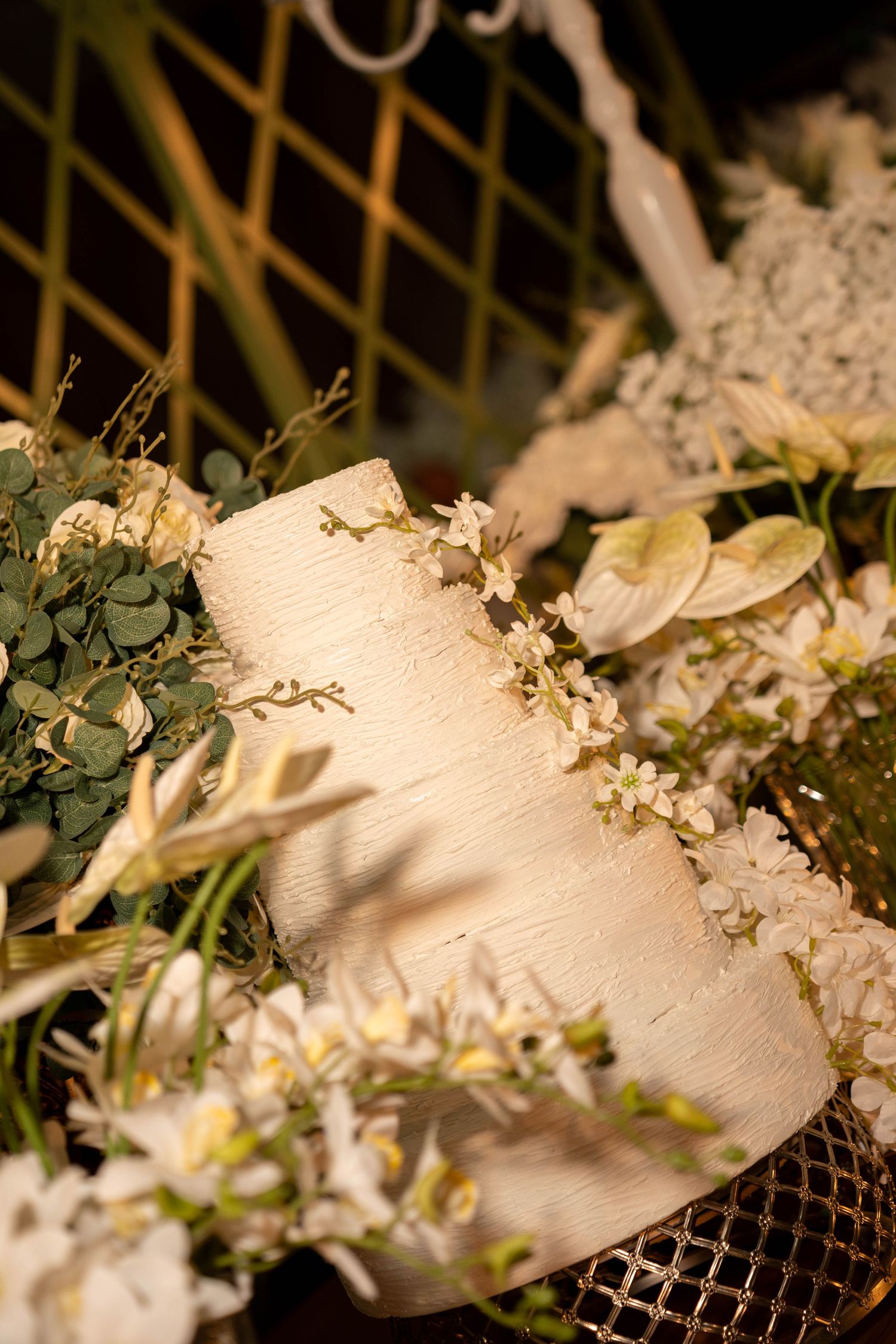 Elegant two-tier white wedding cake adorned with small flowers, surrounded by lush greenery and white floral arrangements.