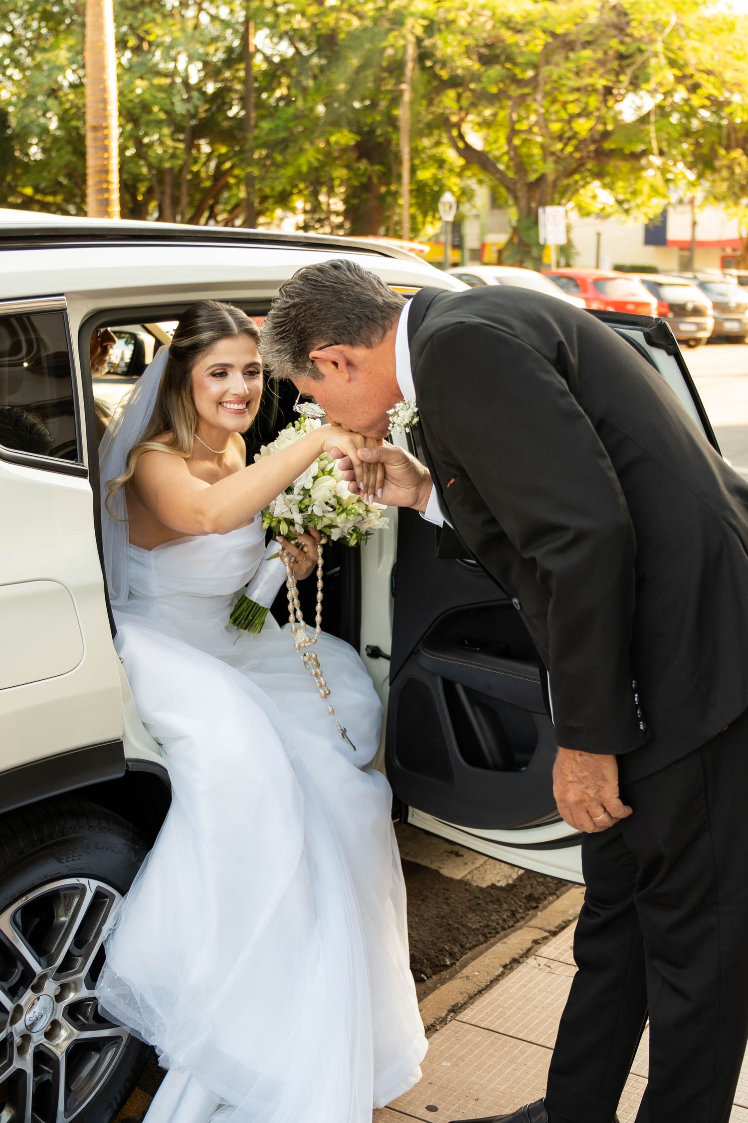 Groom kissing bride's hand as she sits in a white car, holding a bouquet, wearing a wedding dress and veil.