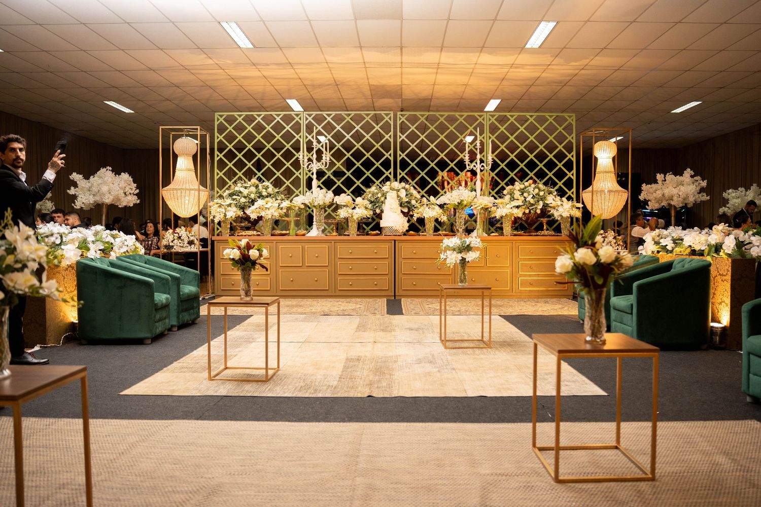 Elegant wedding reception hall with gold accents, green velvet chairs, white floral arrangements, and decorative lattice backdrop.