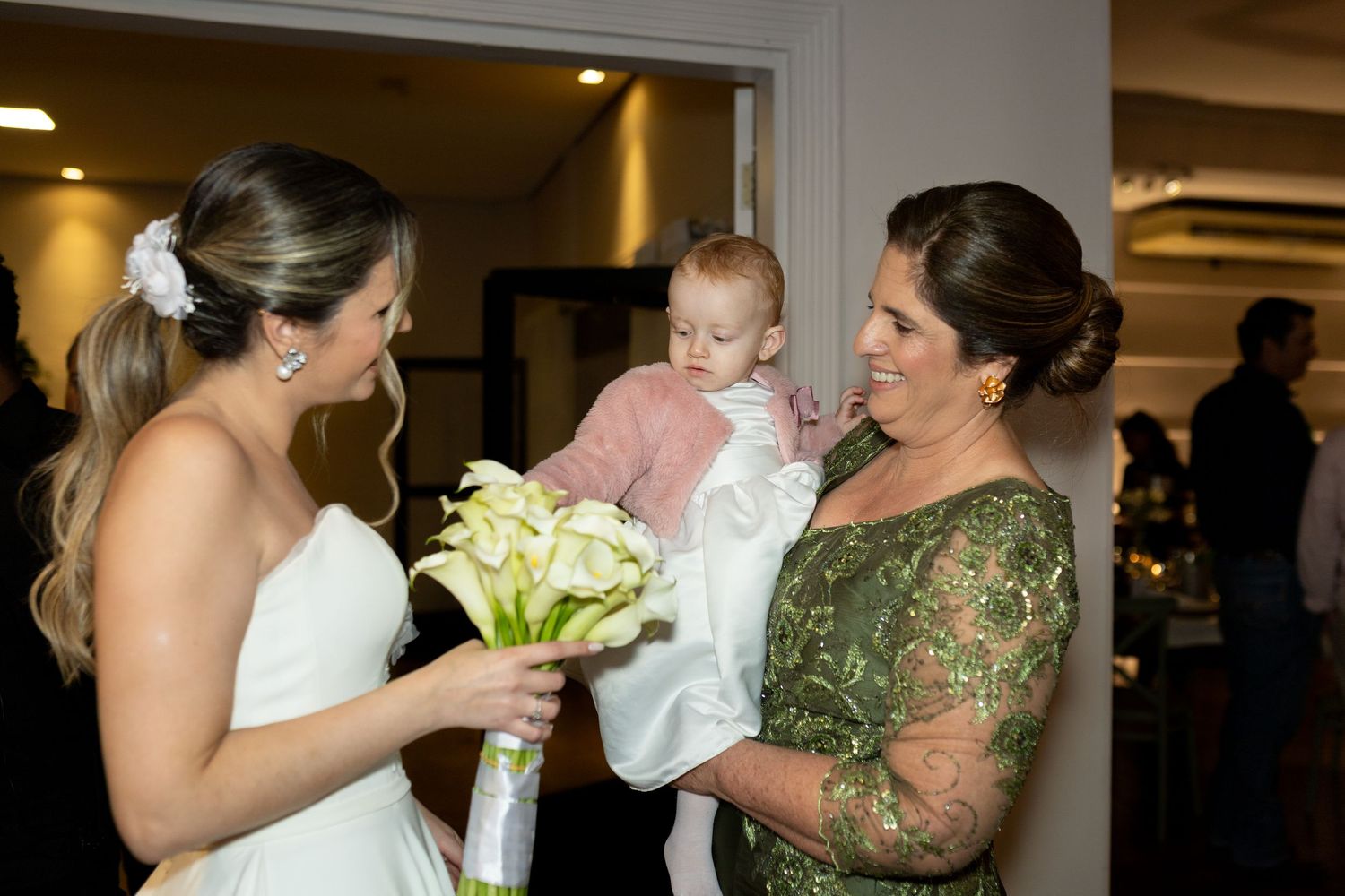 Bride in white gown holds calla lily bouquet while smiling at baby held by woman in green lace dress at wedding reception.
