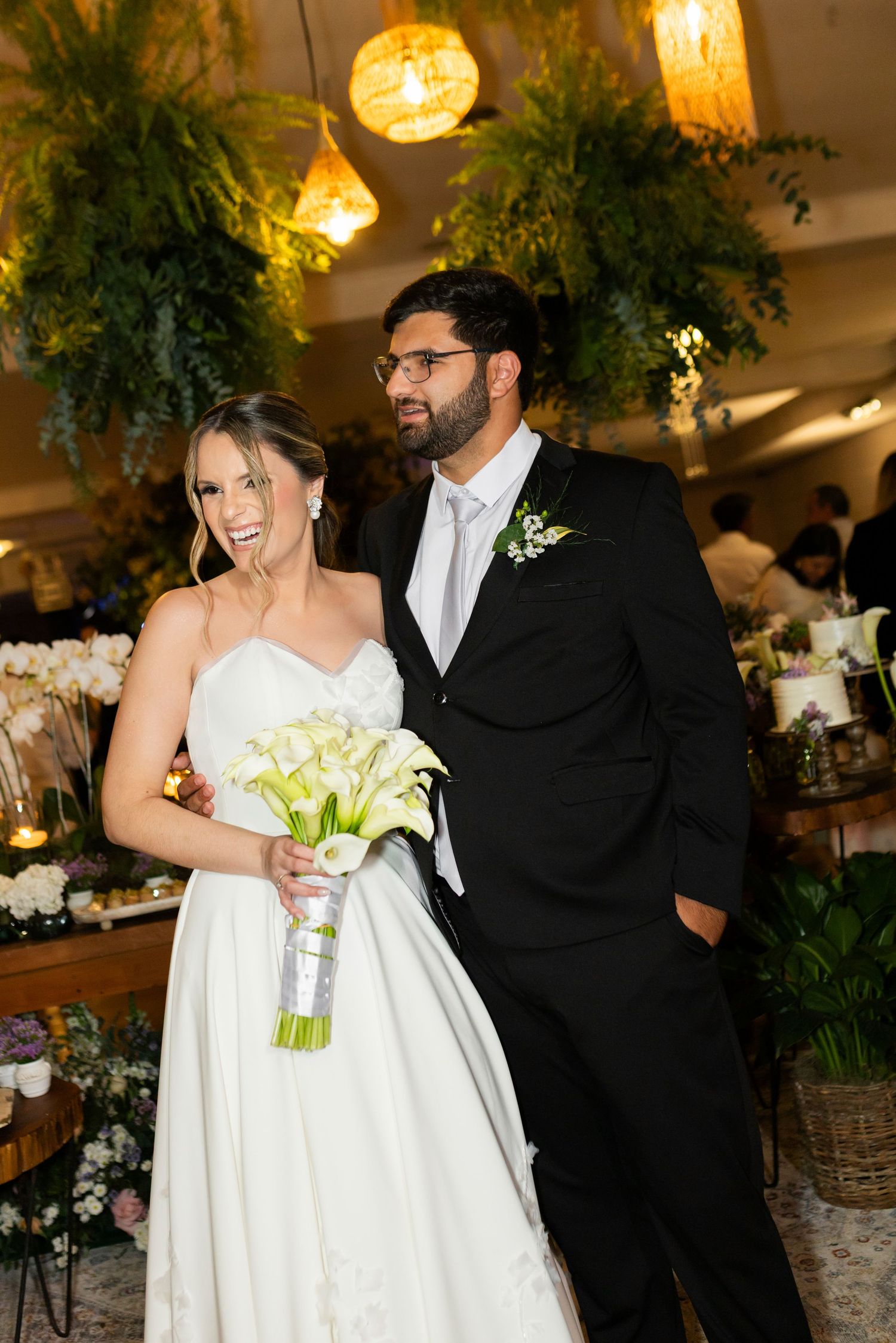 A smiling bride in a white gown holds calla lilies beside a suited groom with boutonniere at an elegant wedding reception.