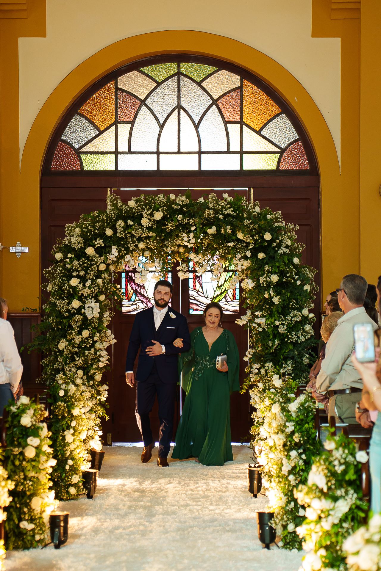 Couple walks down floral arch aisle at elegant church wedding ceremony with white flowers and greenery.