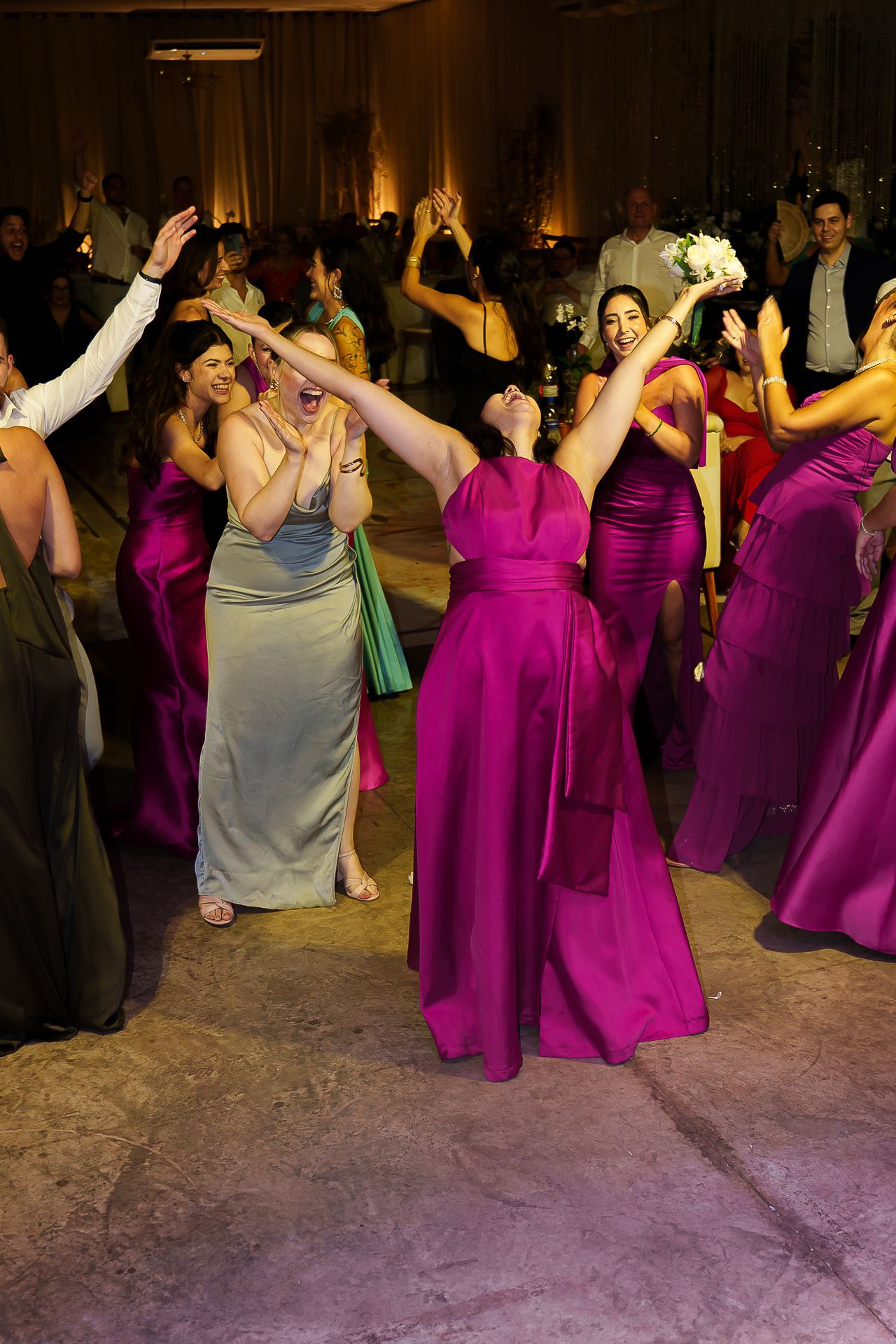 Bridesmaids in magenta and satin gowns dancing joyfully at a wedding reception.
