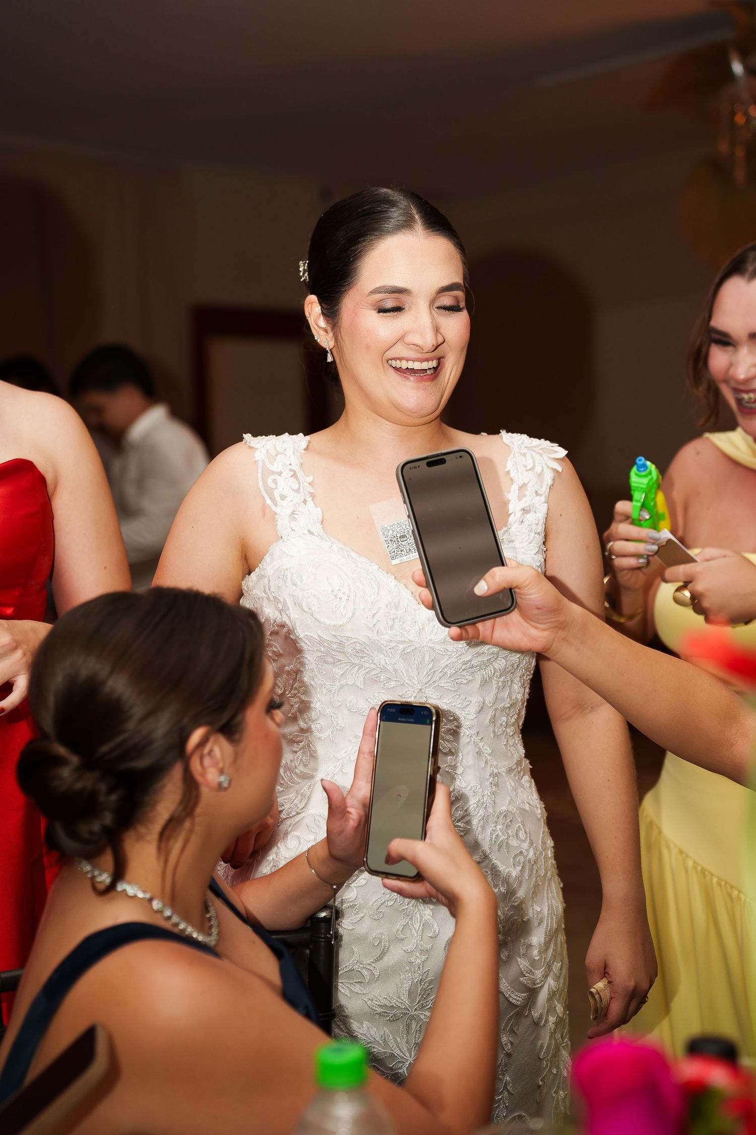Smiling bride in white lace gown surrounded by guests showing phones at a wedding reception celebration.