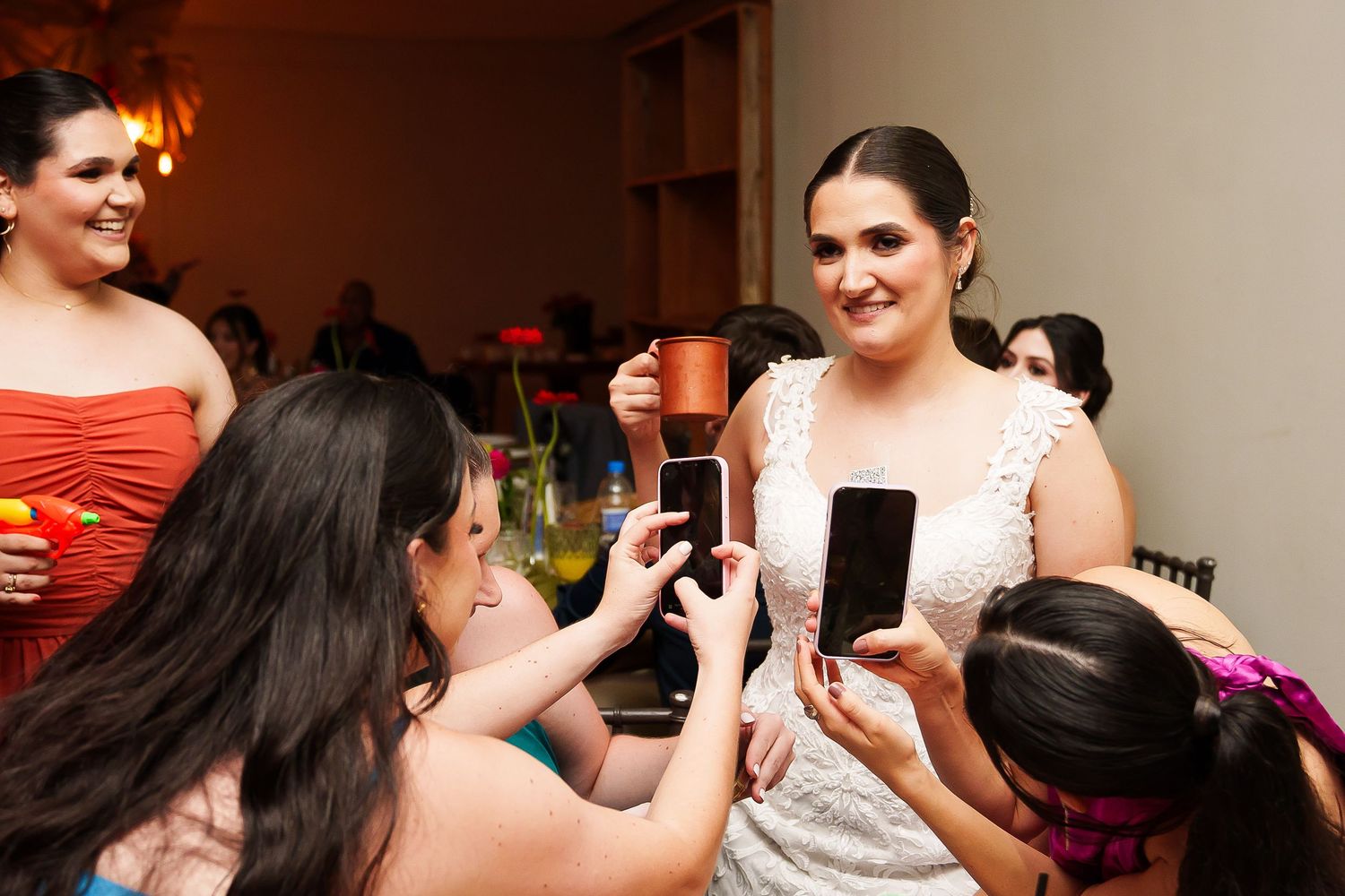 Bride in white lace dress smiles as guests photograph her with smartphones at a wedding reception.