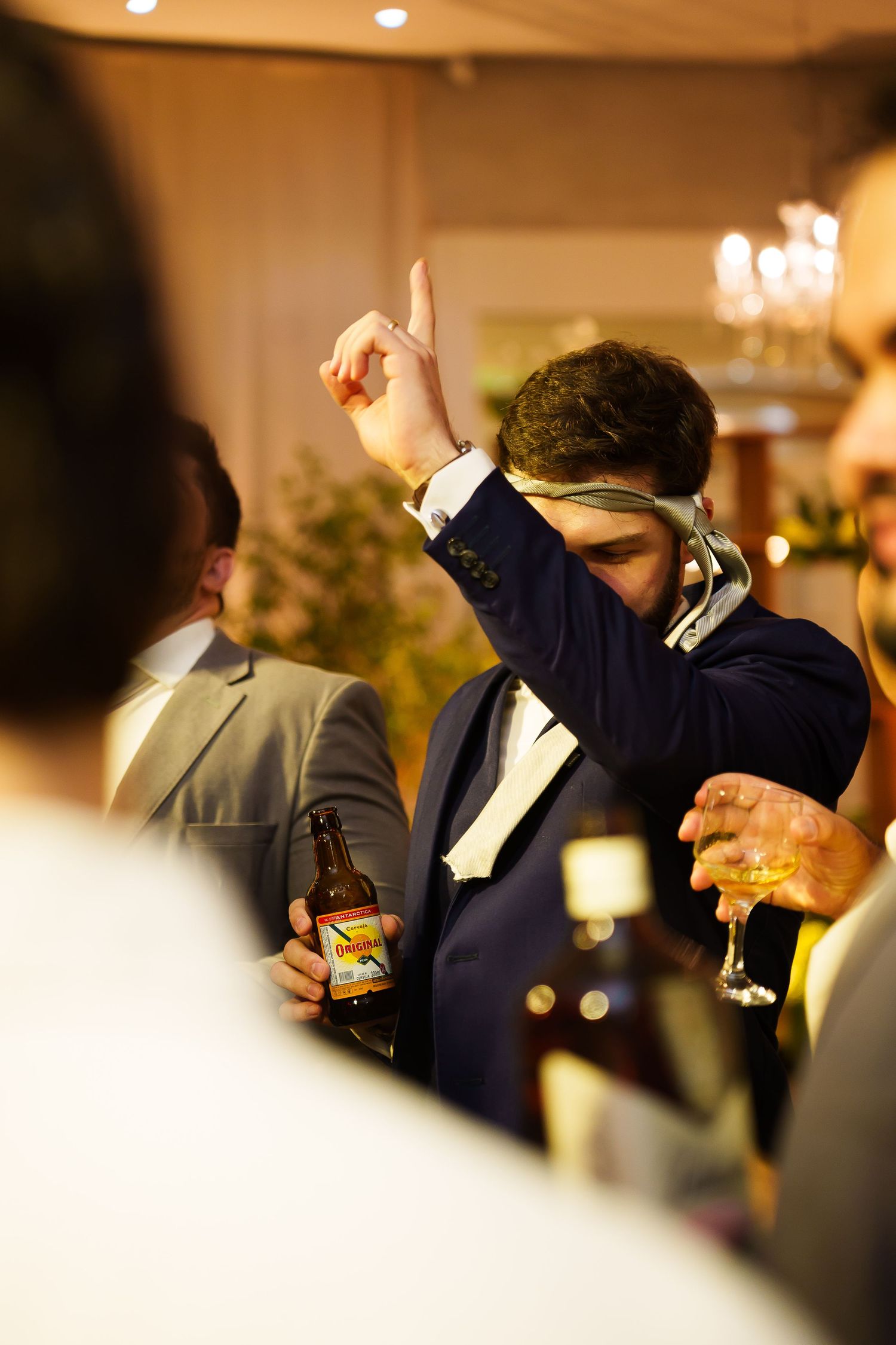 Man in suit dancing at wedding reception, holding beer bottle and glass while wearing sunglasses indoors.