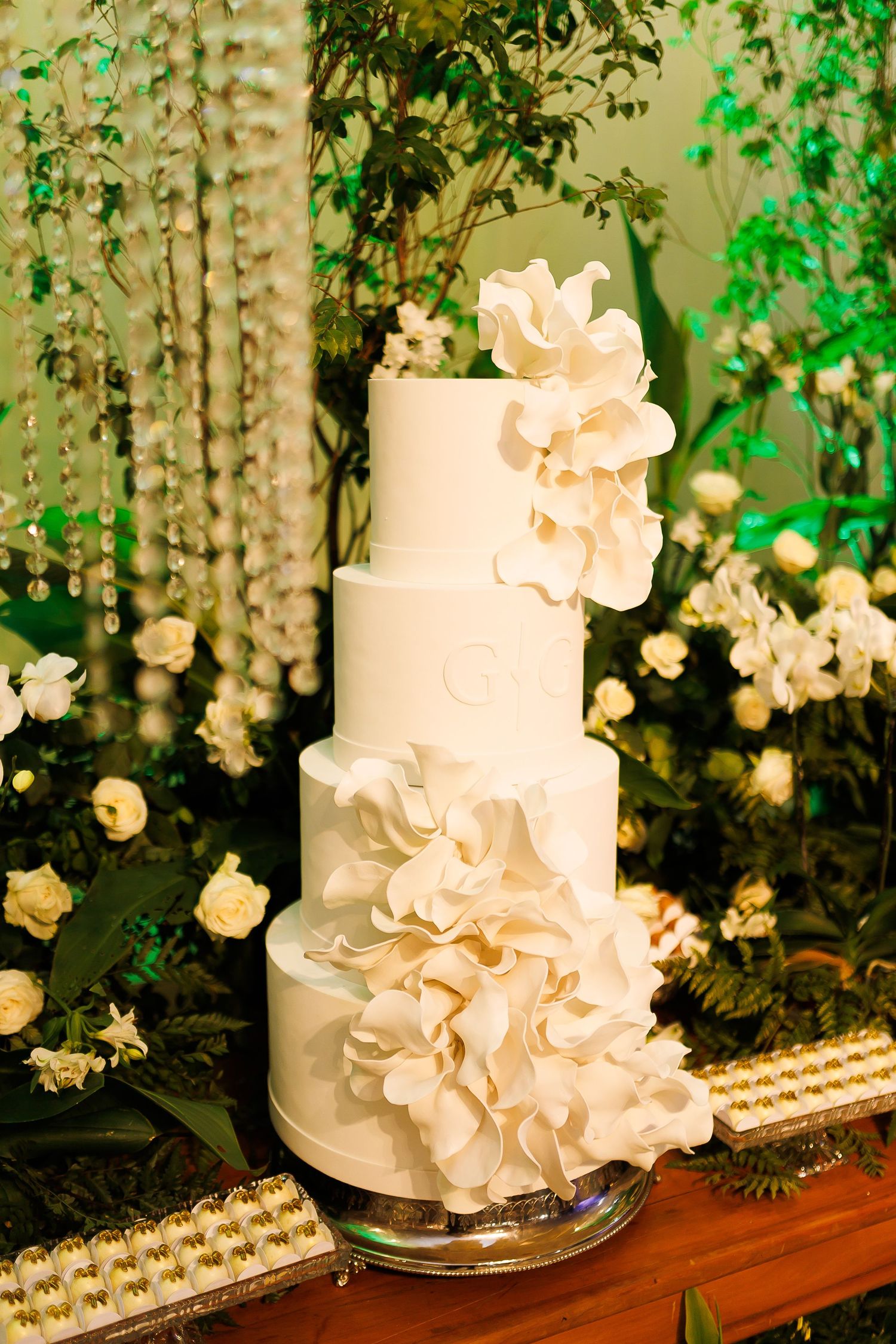 Elegant four-tier white wedding cake adorned with large sugar flowers, displayed against a lush green floral backdrop.