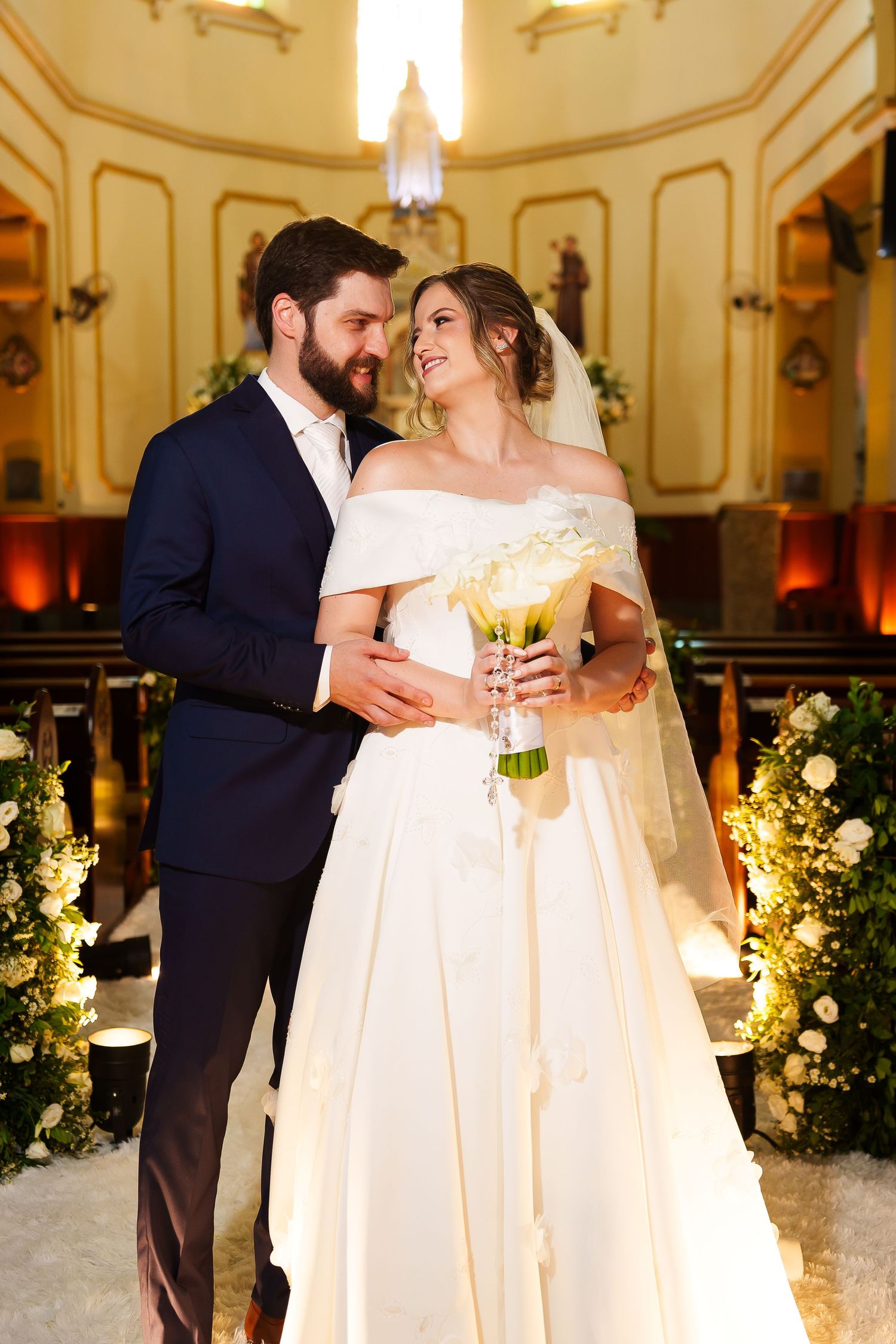 Bride and groom embrace in church aisle, bride holds white bouquet in elegant off-shoulder gown with veil.