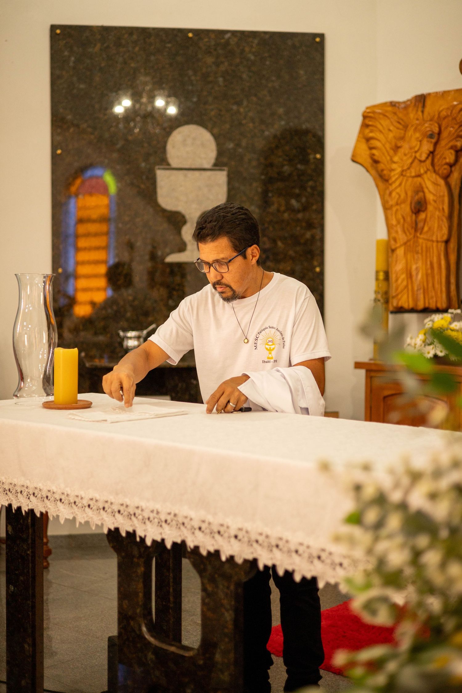 A priest in white vestments stands at an altar reviewing documents in a church setting.