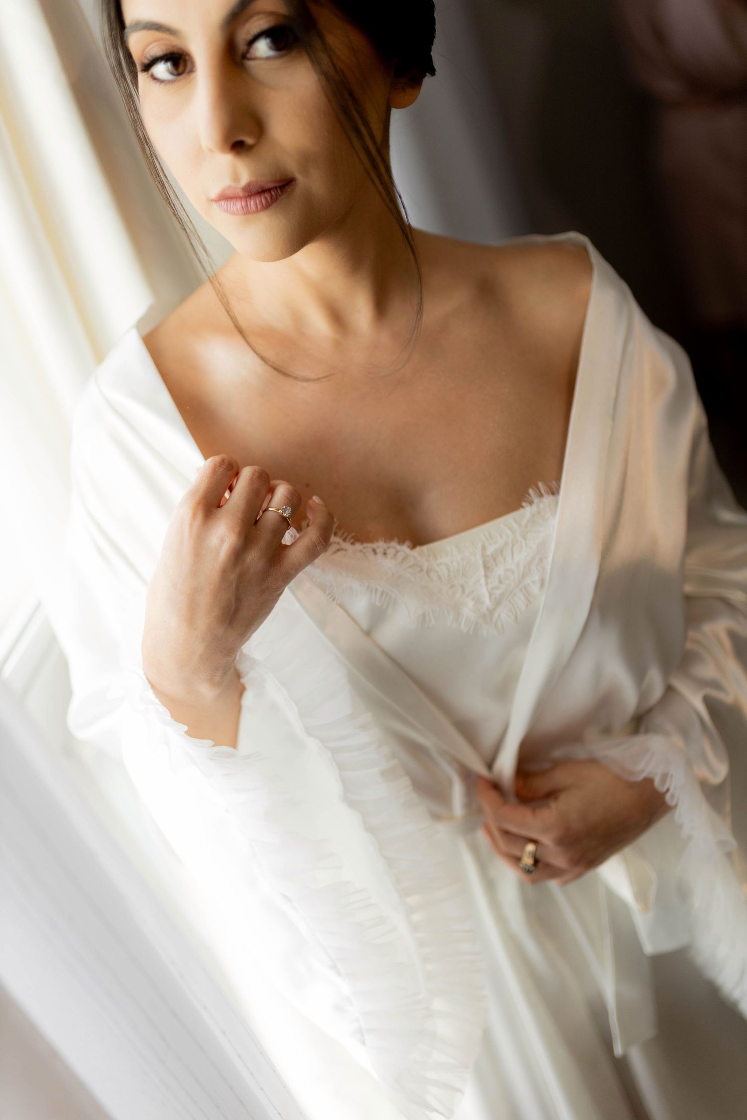 A bride in a white silk robe and lace lingerie gazes thoughtfully toward a window, adorned with elegant gold rings.