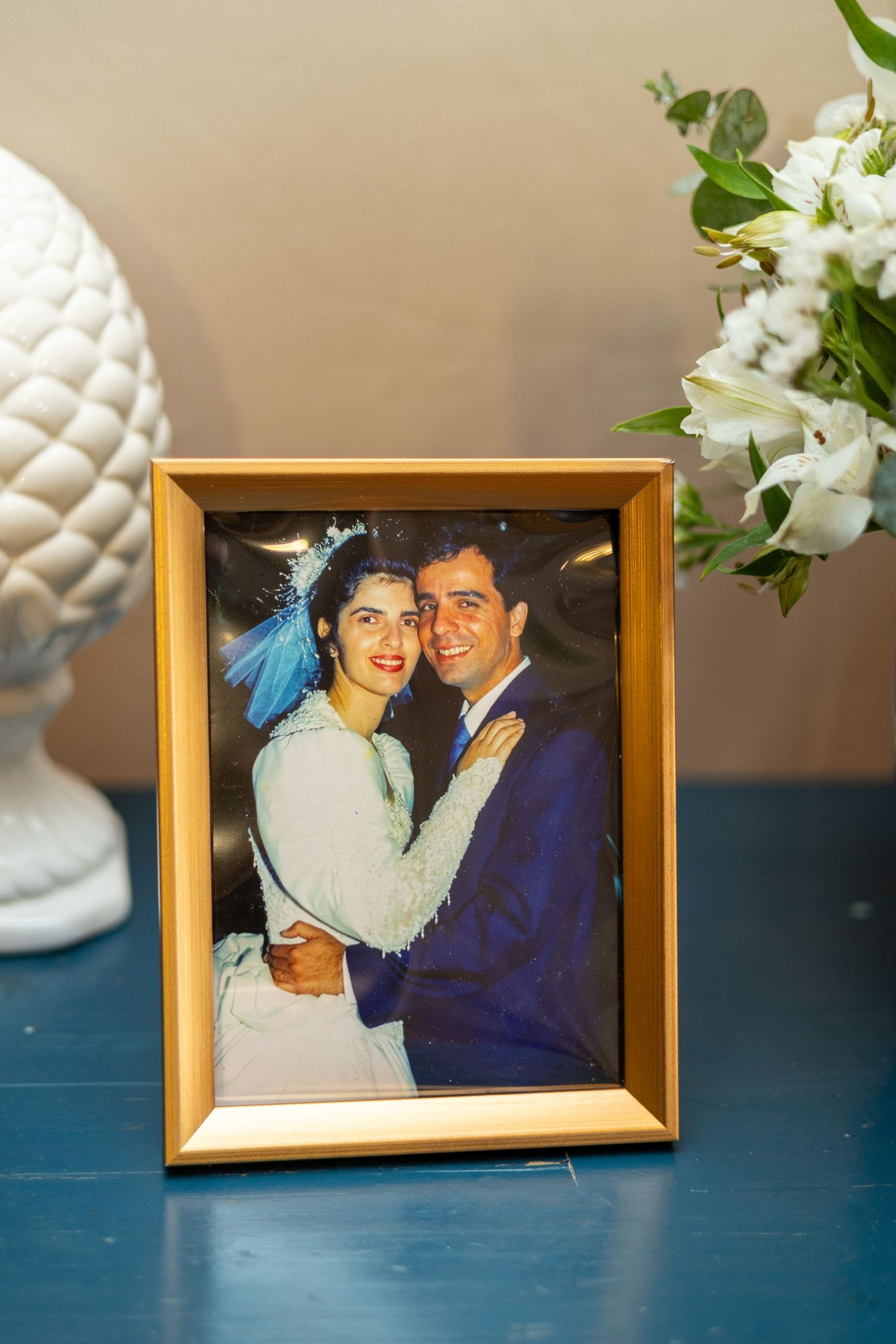 Framed wedding photo of a smiling couple in gold frame on blue table with white flowers and decorative pine cone.