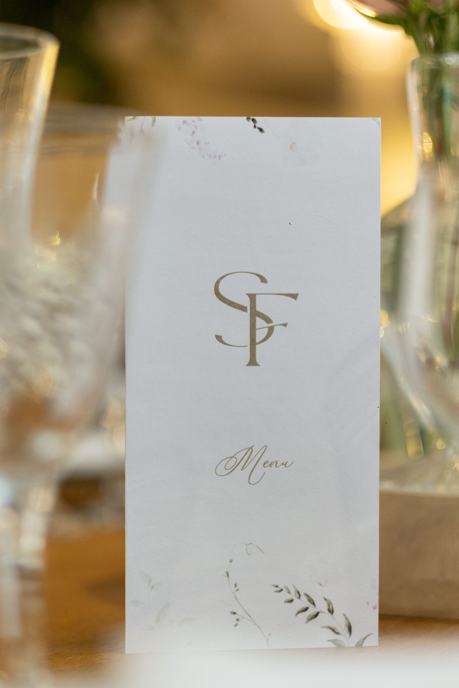 Elegant white wedding menu card with gold SF monogram and delicate botanical details on a reception table.