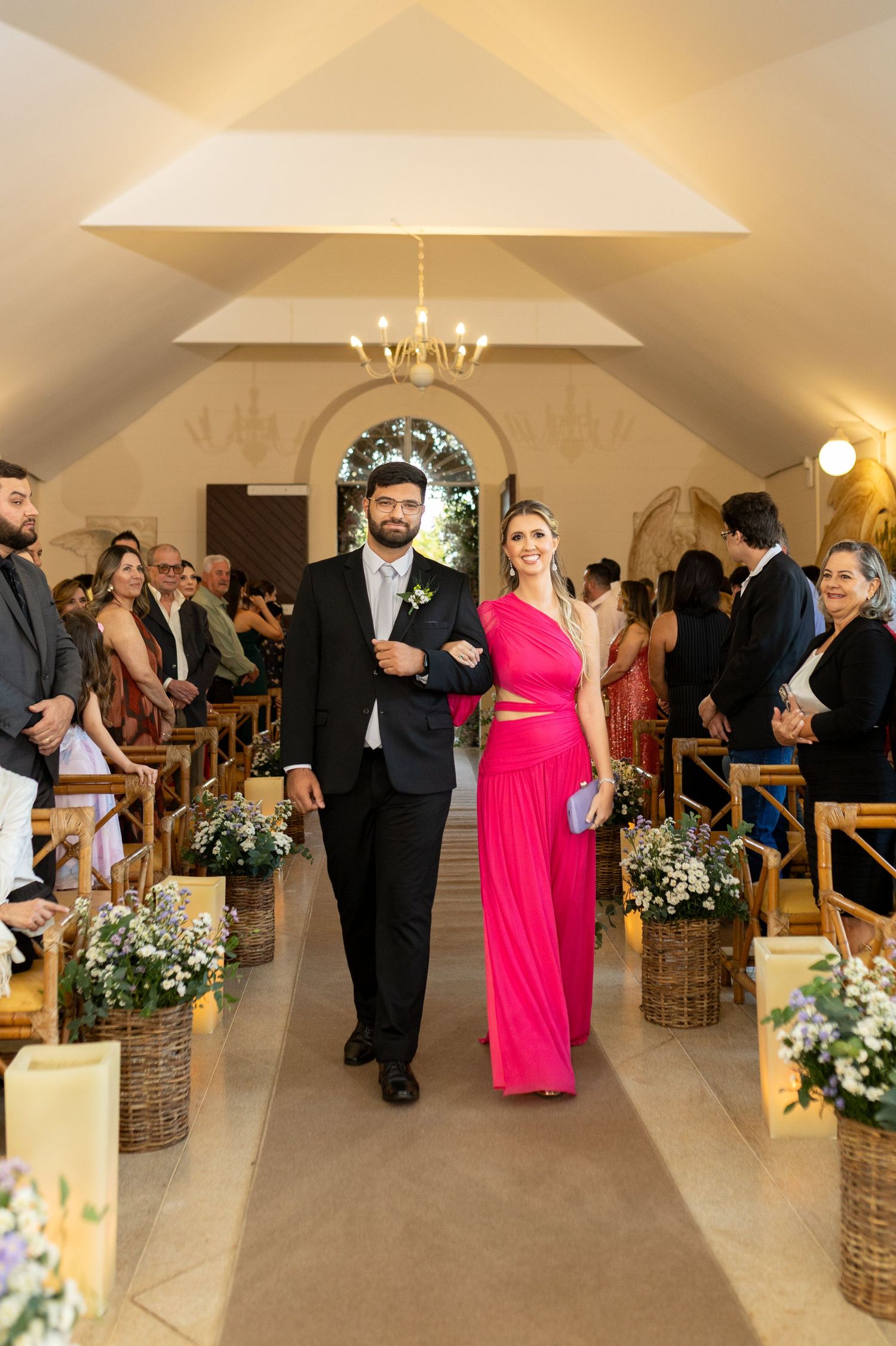 A couple walks down a church aisle, man in dark suit, woman in bright pink gown, flanked by floral arrangements.