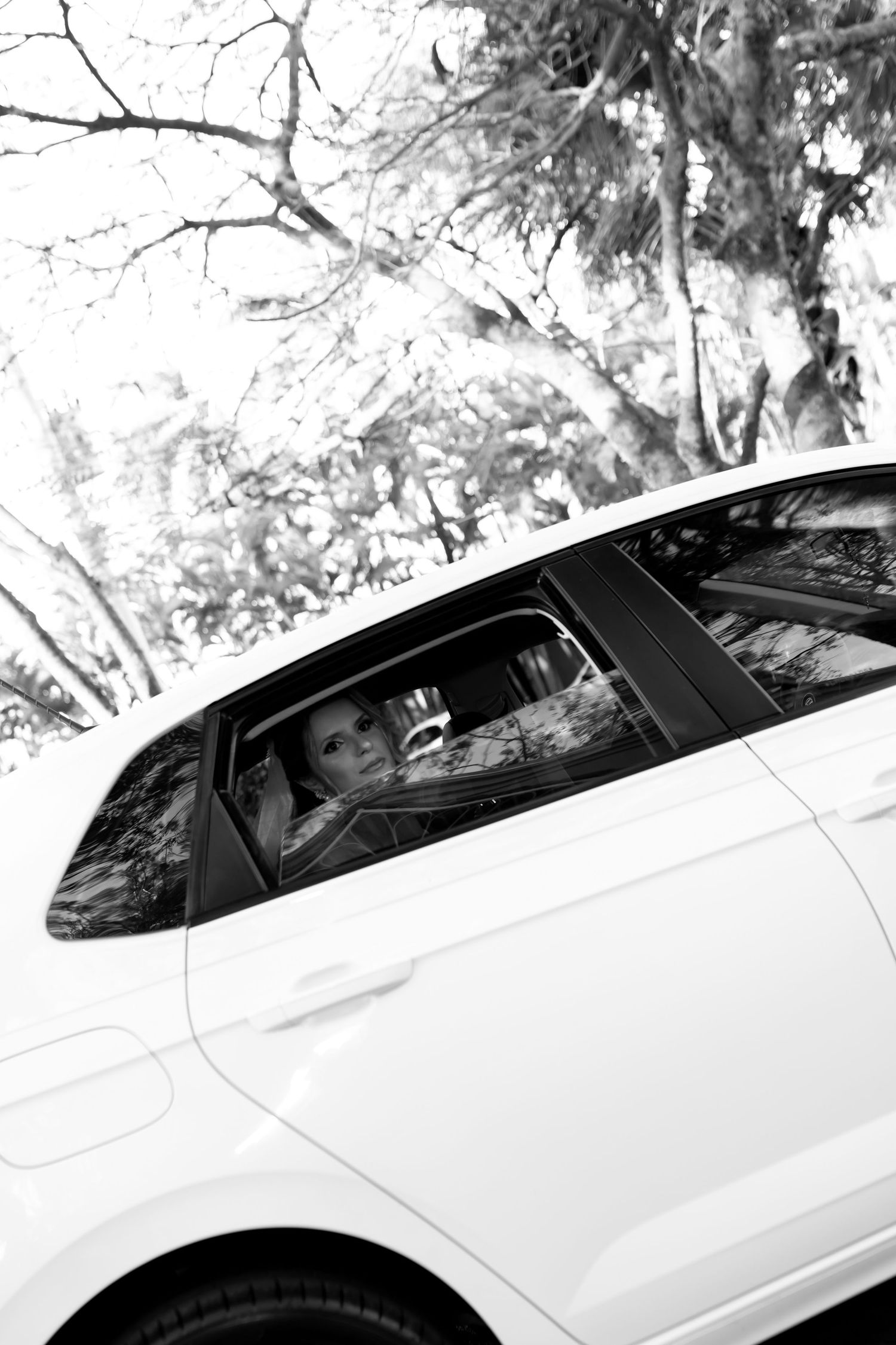 Black and white photo of a woman sitting in a white car, surrounded by trees, looking out the window.