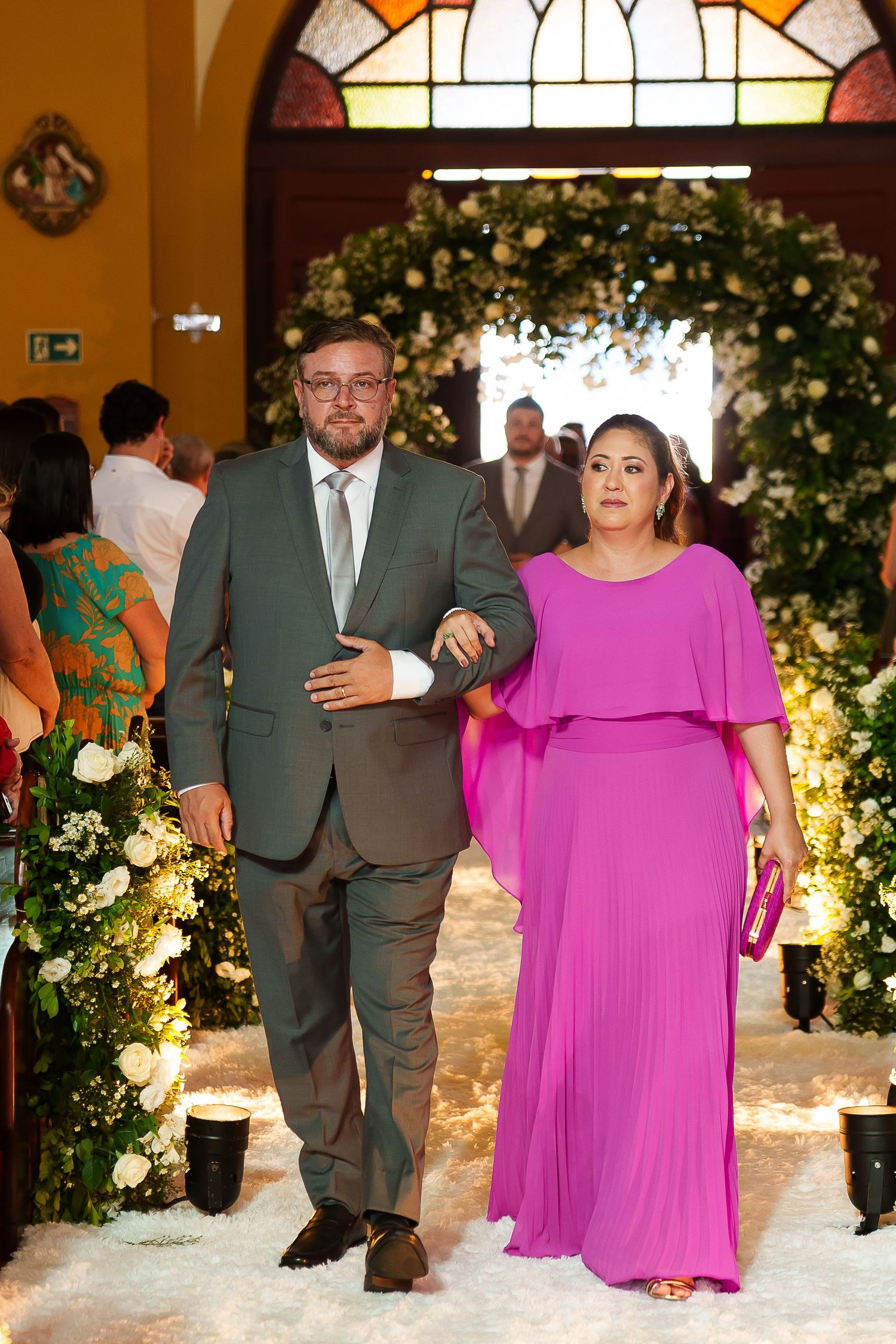 Couple walking down wedding aisle, man in gray suit, woman in pink gown, floral decorations.