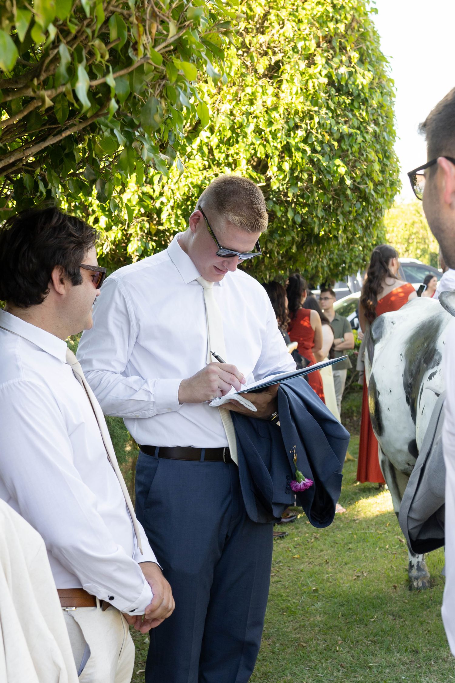 Man in white shirt writing on clipboard outdoors at a garden event with guests in background.