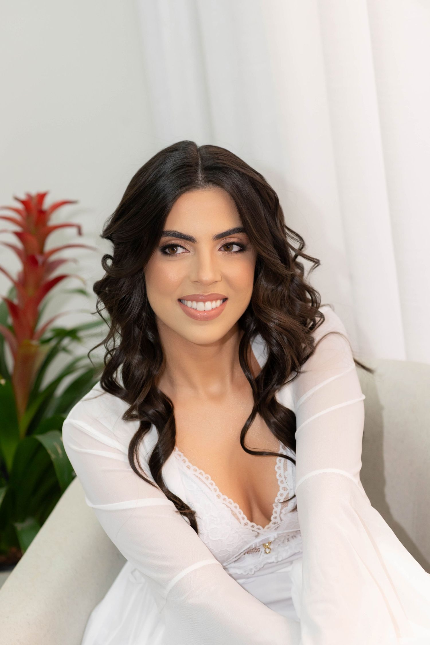 Smiling brunette bride in white lace gown with sheer sleeves, posing elegantly in a bright room with tropical plants.
