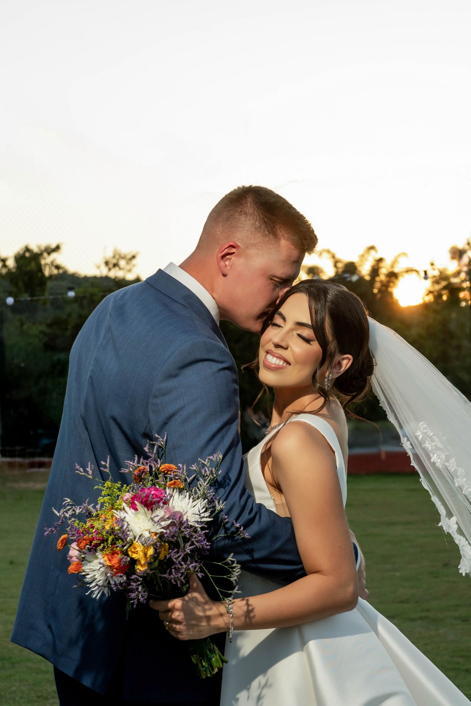 Bride and groom share a romantic sunset moment, she holds a colorful wildflower bouquet while he kisses her cheek.