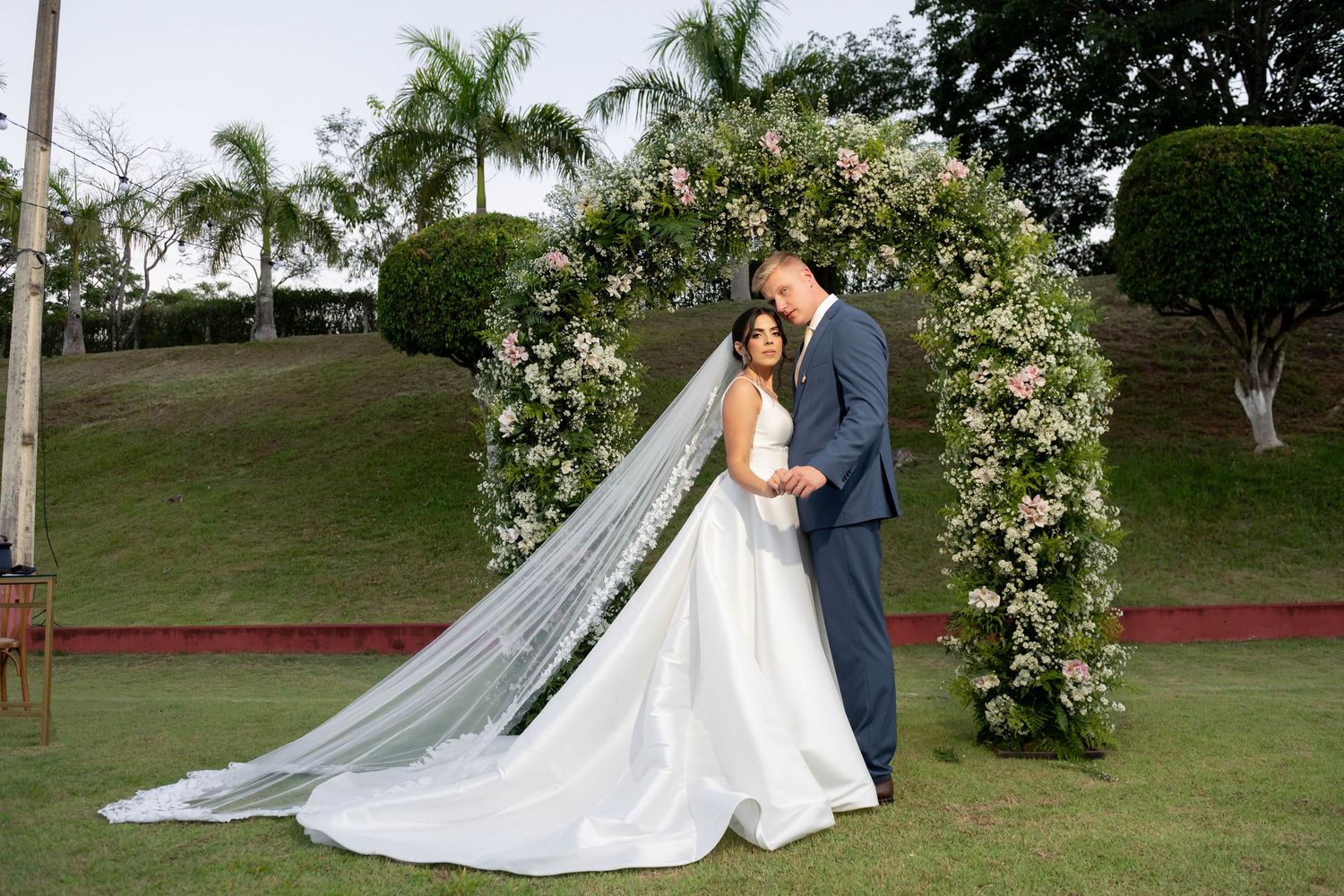 Bride and groom share a romantic moment under a floral arch on a lush green lawn with palm trees.