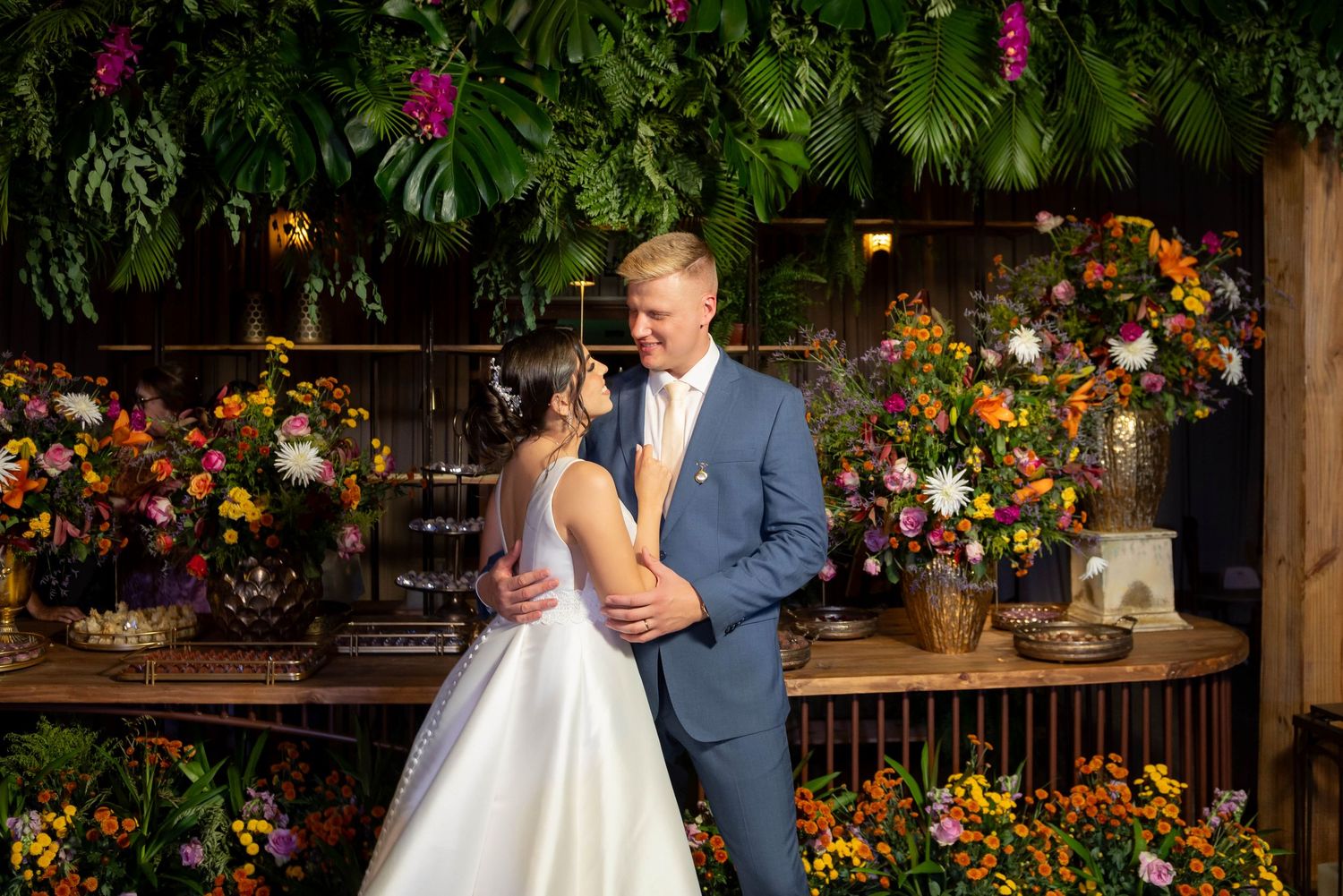 Bride and groom embracing in front of lush tropical greenery and colorful floral arrangements at a wedding reception.