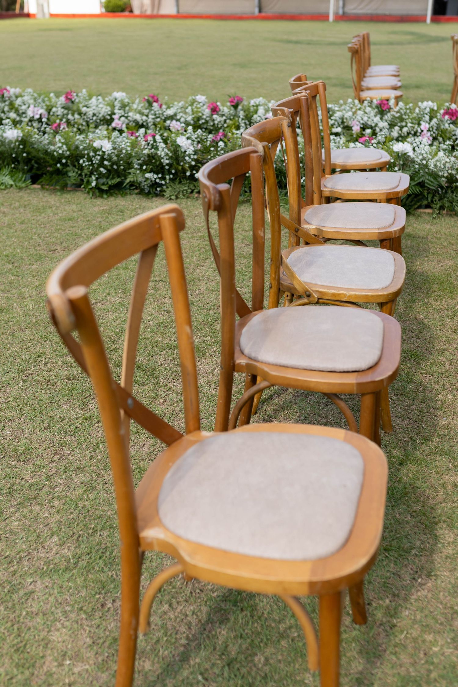 Row of wooden cross-back chairs with cushioned seats arranged on green grass near a floral garden for an outdoor event.