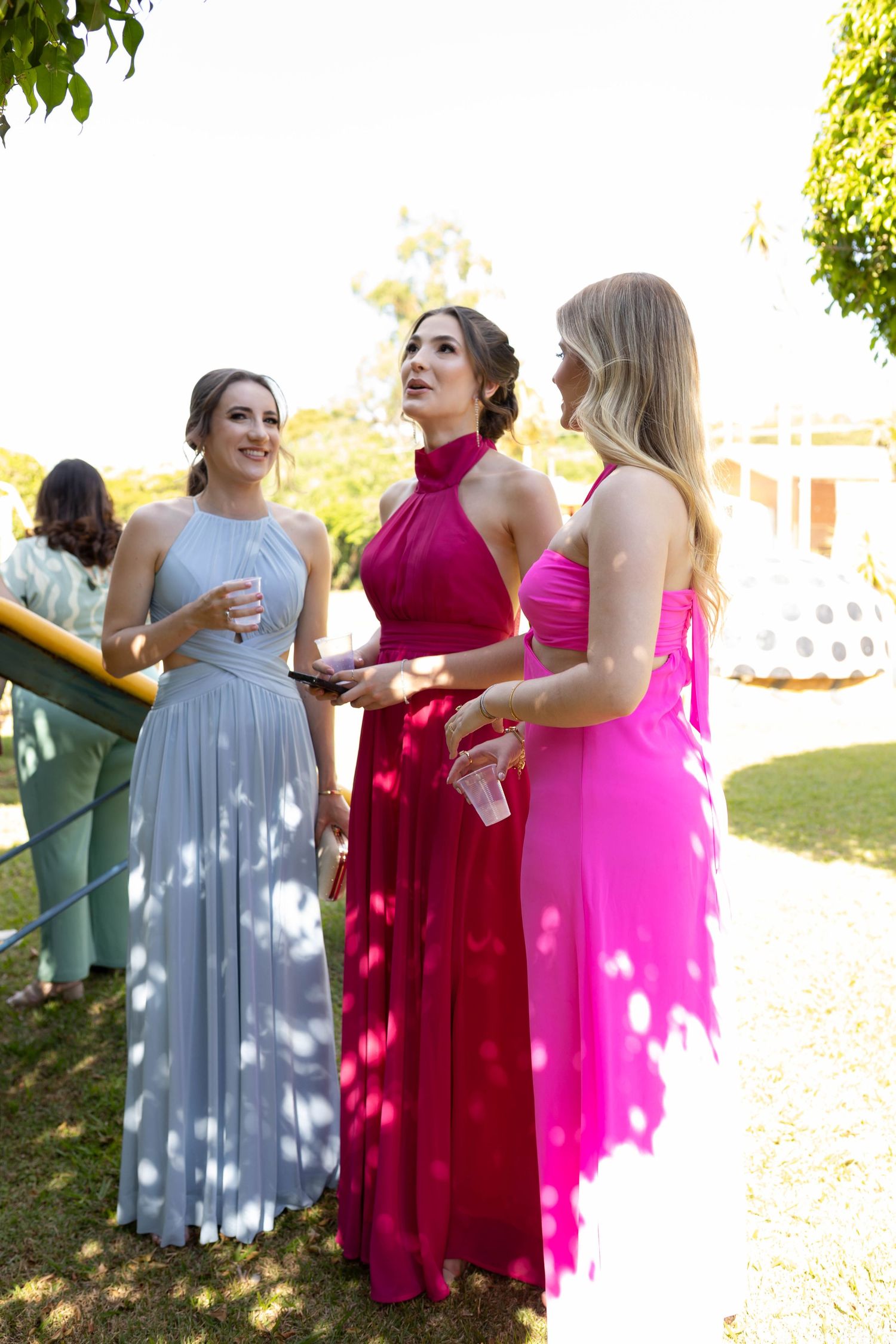 Three women in elegant gowns of silver, magenta, and pink chat outdoors at a formal garden event.