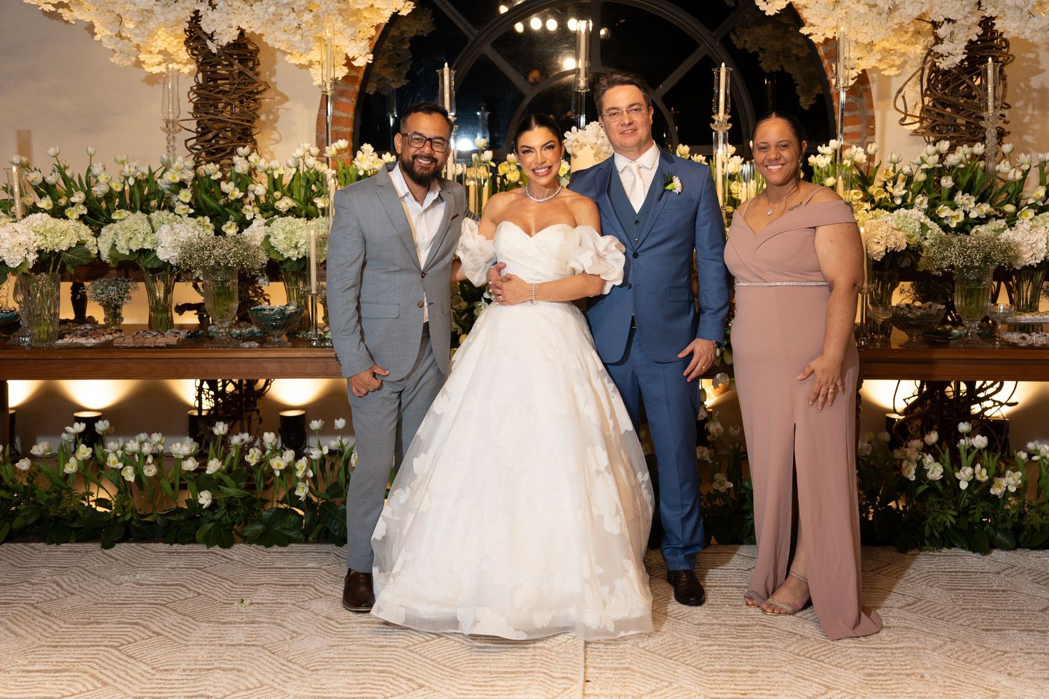 Bride in white ball gown poses with groom and guests at elegant floral wedding reception.