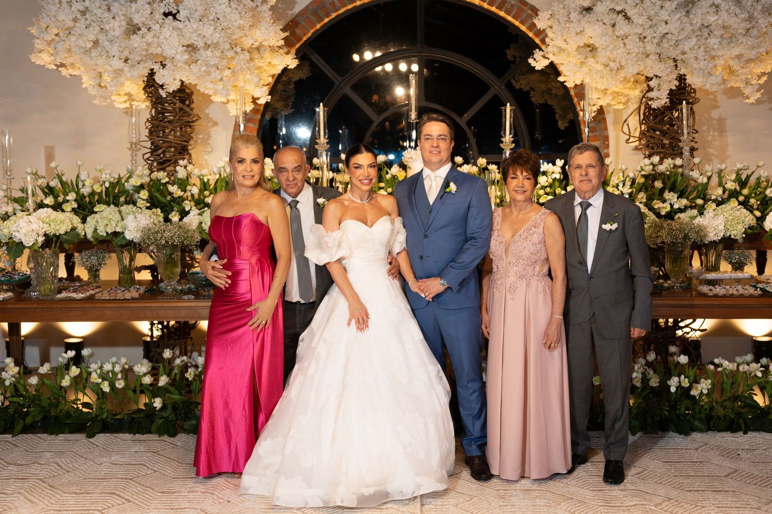 Wedding party of five posing elegantly at a floral-decorated reception venue, featuring bride in white gown and groom in blue suit.