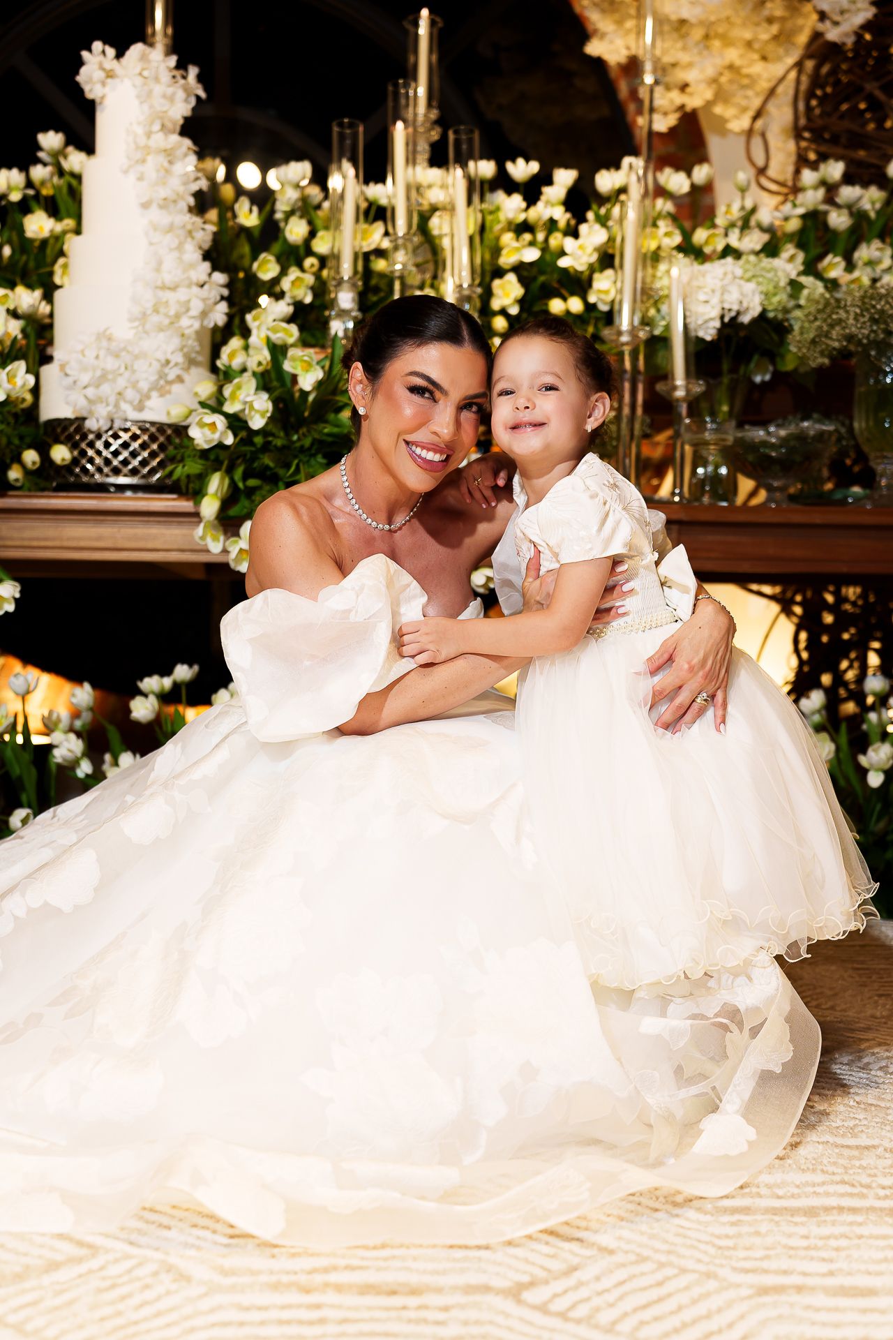 Bride in white ball gown hugs young flower girl in white dress, posing together at elegant wedding reception.