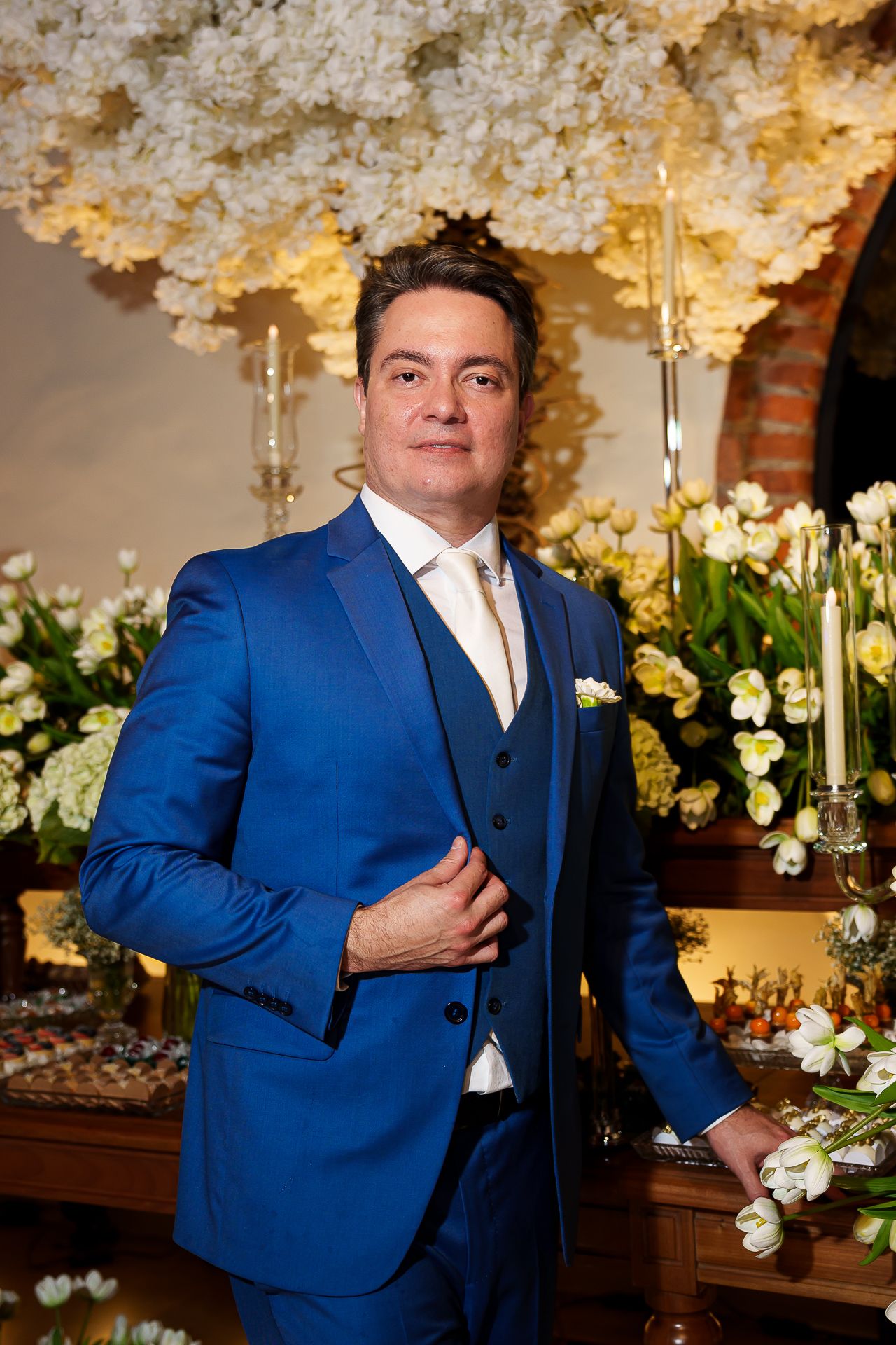 Elegant man in blue three-piece suit posing at floral wedding reception with white flower arrangements.
