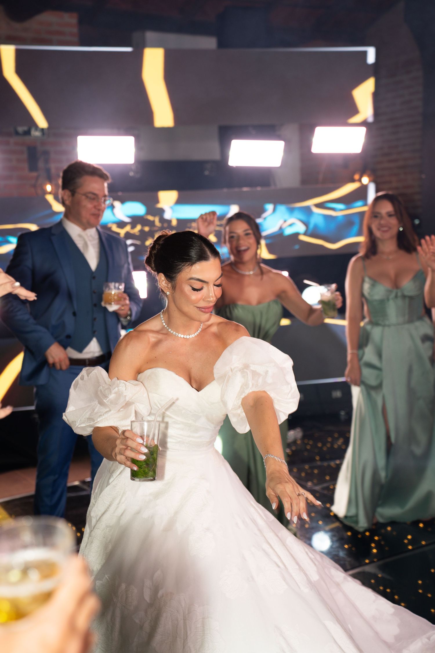 Bride in white gown holds cocktail on dance floor, surrounded by guests in blue and green attire at wedding reception.