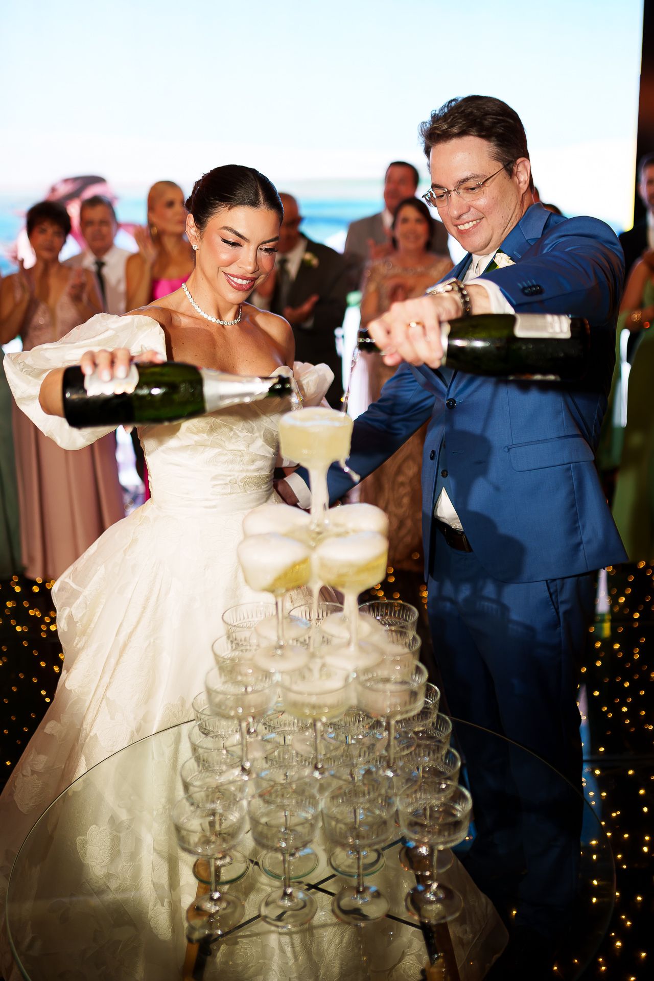 Bride and groom pouring champagne tower at wedding reception, guests watching in background.