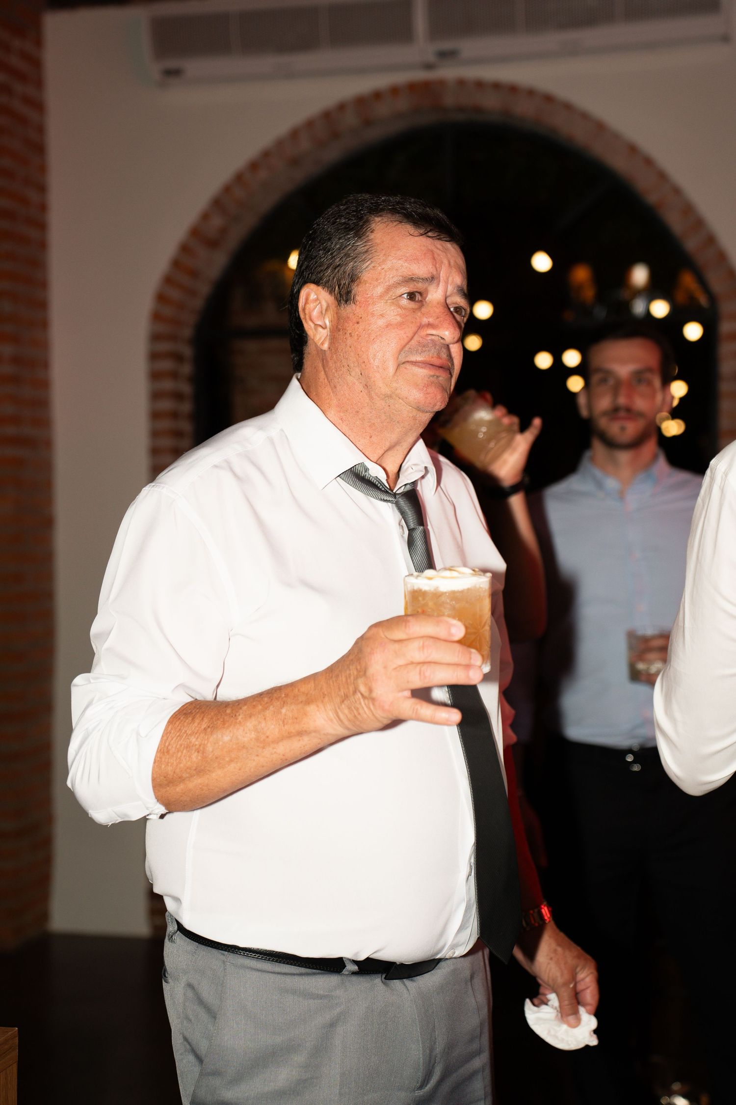 Man in white shirt and tie holding a drink at an evening social event or wedding reception.
