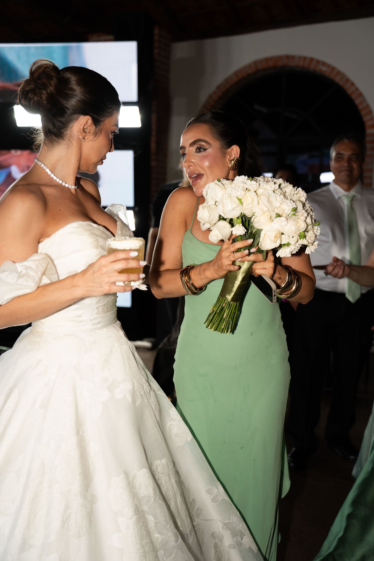 Bride in white gown holds drink while bridesmaid in green dress reacts with surprise holding white rose bouquet.