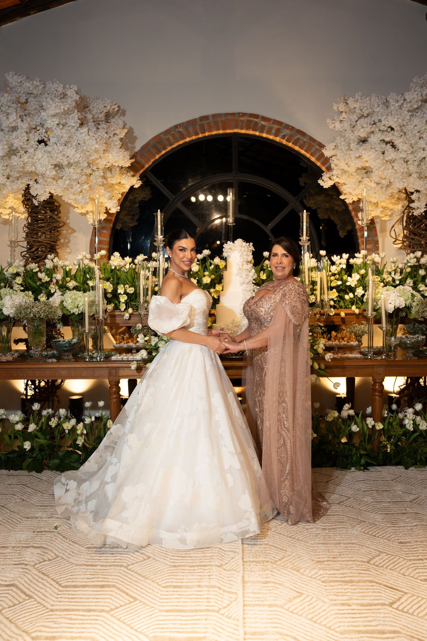 Bride in white ball gown and woman in gold dress cutting a tiered wedding cake surrounded by white floral arrangements.
