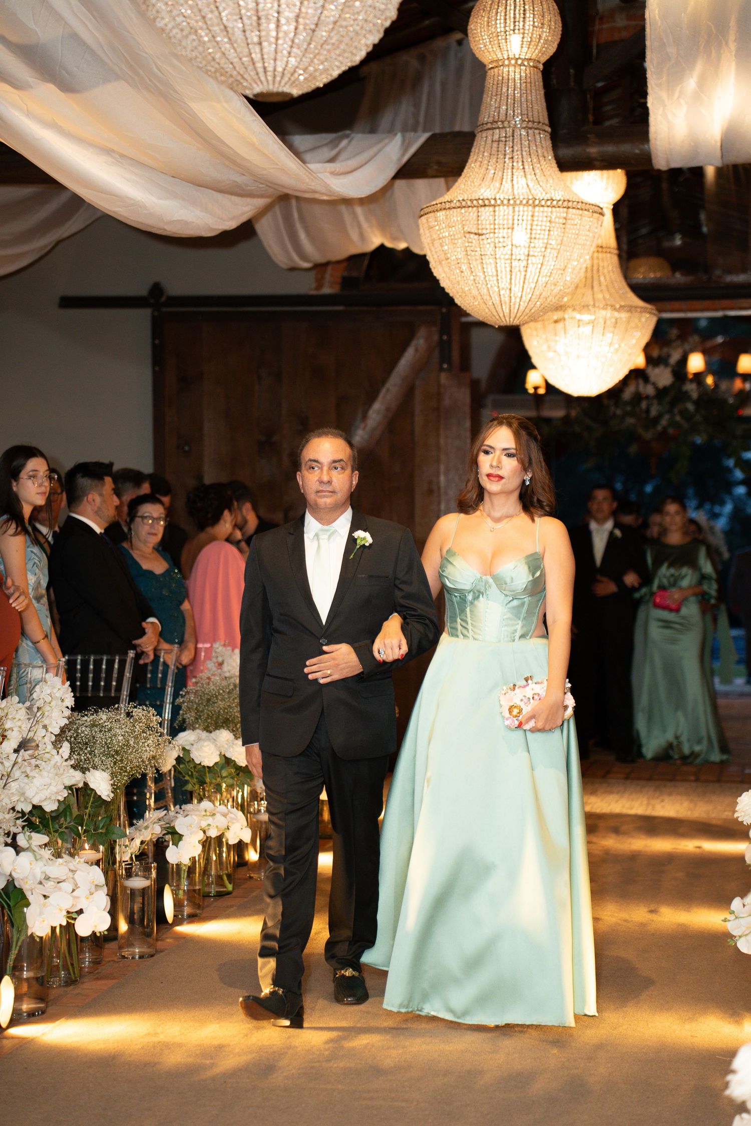 Elegant couple walks down wedding aisle, woman in mint green gown, man in black tuxedo, chandeliers above.