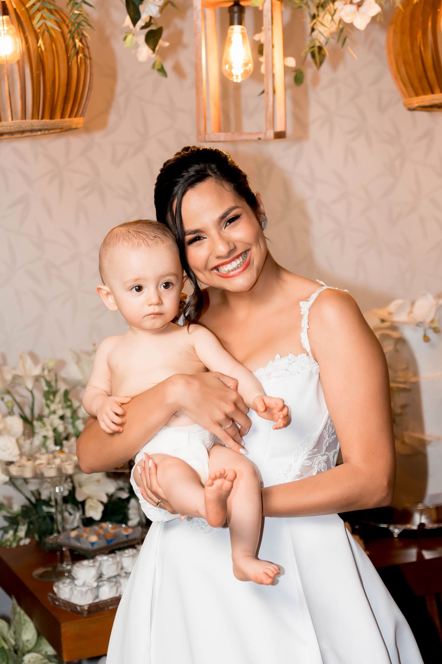 Smiling bride in white lace gown holds a baby at a beautifully decorated wedding reception.