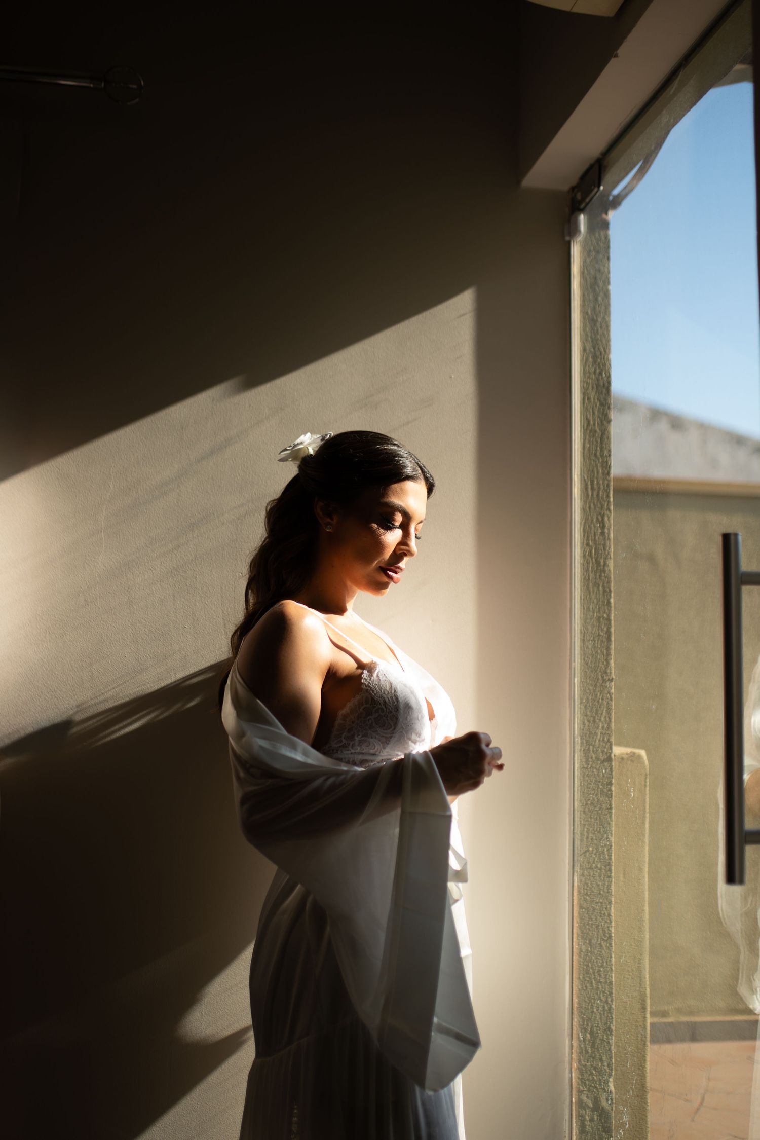 Bride in white wedding dress stands by sunlit window, soft light casting dramatic shadows on the wall.