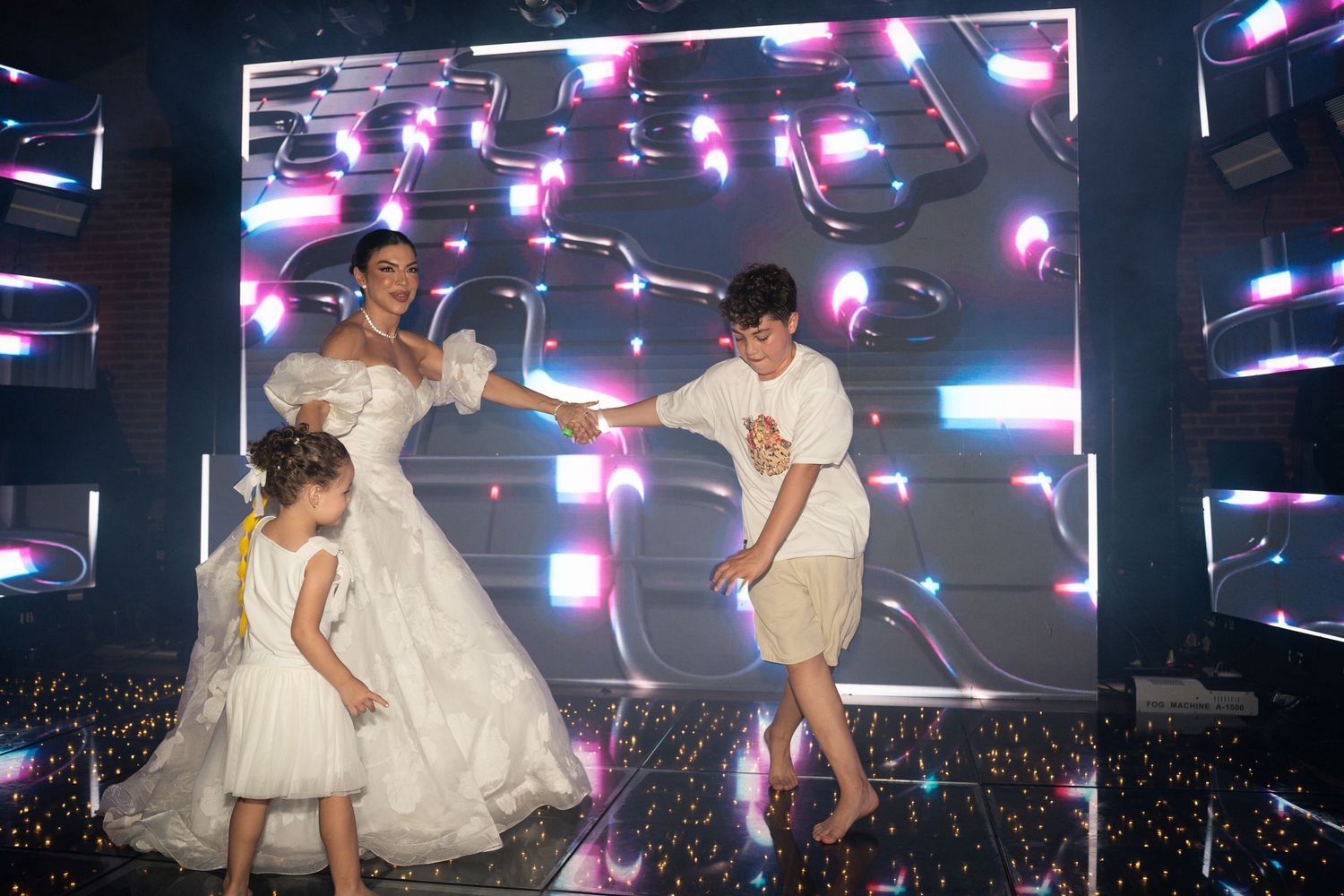 Woman in white gown dances on stage with two children in front of a colorful LED display backdrop.