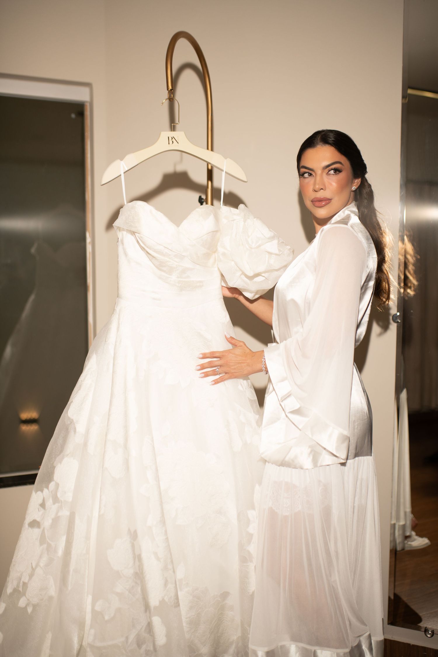 Bride in white robe admiring elegant strapless wedding gown on hanger in bridal boutique fitting room.