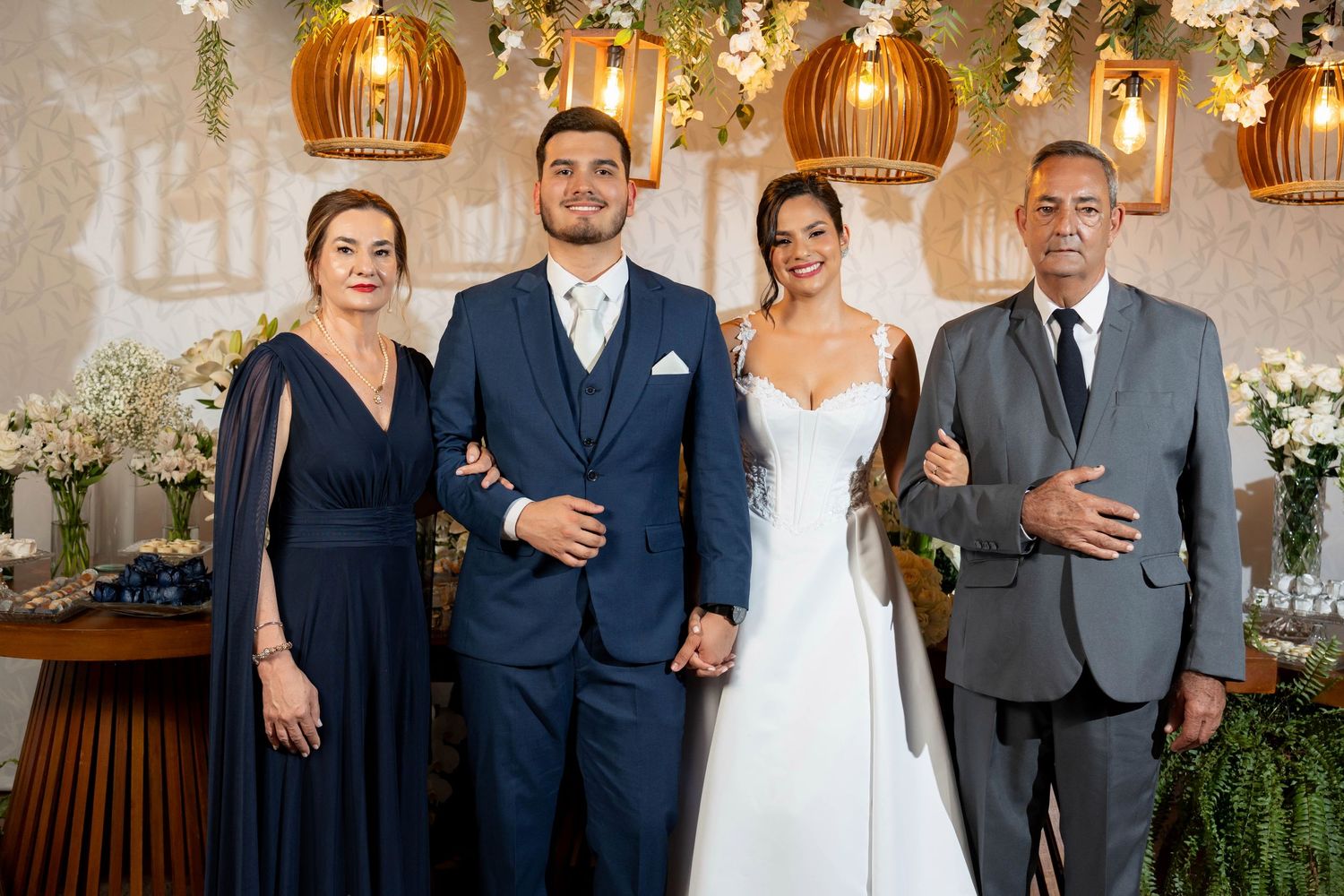Elegant wedding photo with bride in white gown and groom in navy suit posing with family amid floral decor.