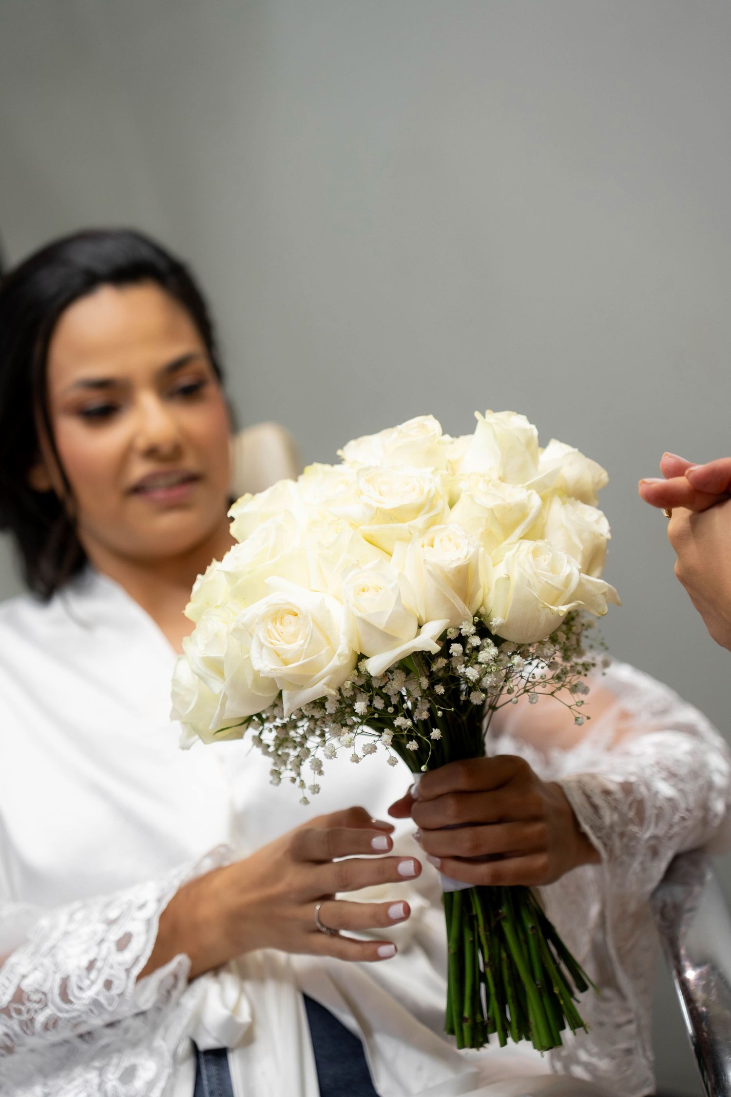 Bride in white lace robe receiving a bouquet of white roses with baby's breath on her wedding day.