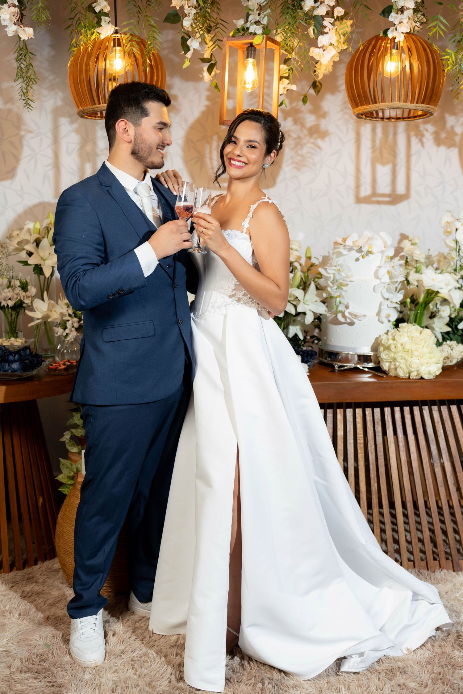 Happy newlyweds toasting with champagne glasses in elegant wedding venue with floral decor and cake.