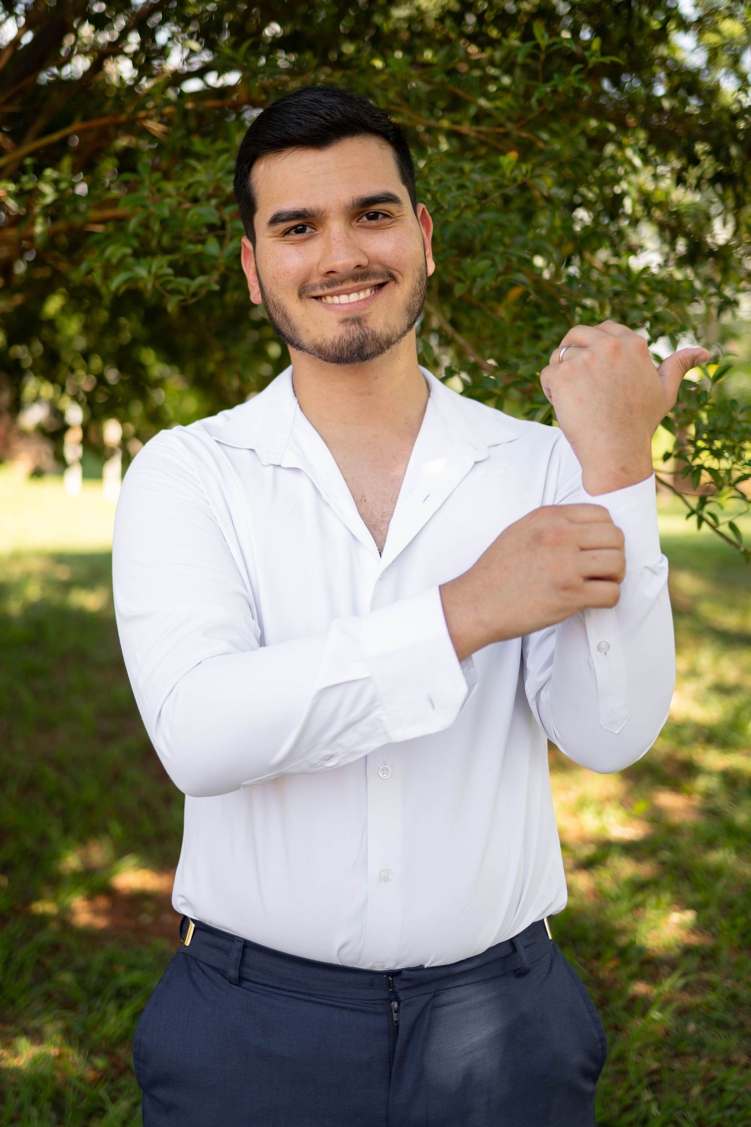 Young man in white dress shirt adjusting cuffs outdoors, smiling confidently with green trees in background.