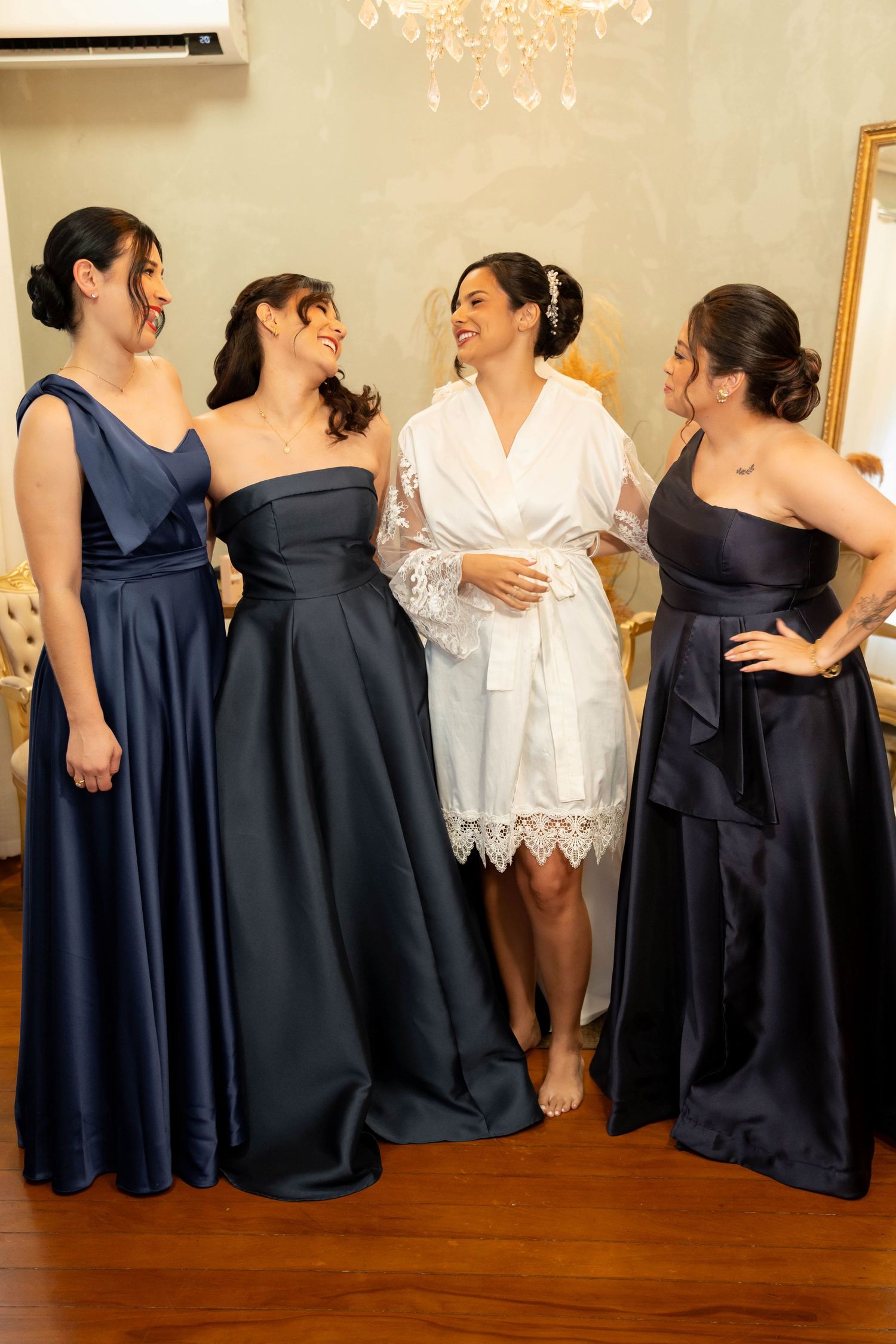 Bride in white lace robe stands with three bridesmaids in navy satin gowns, smiling together in an elegant room.