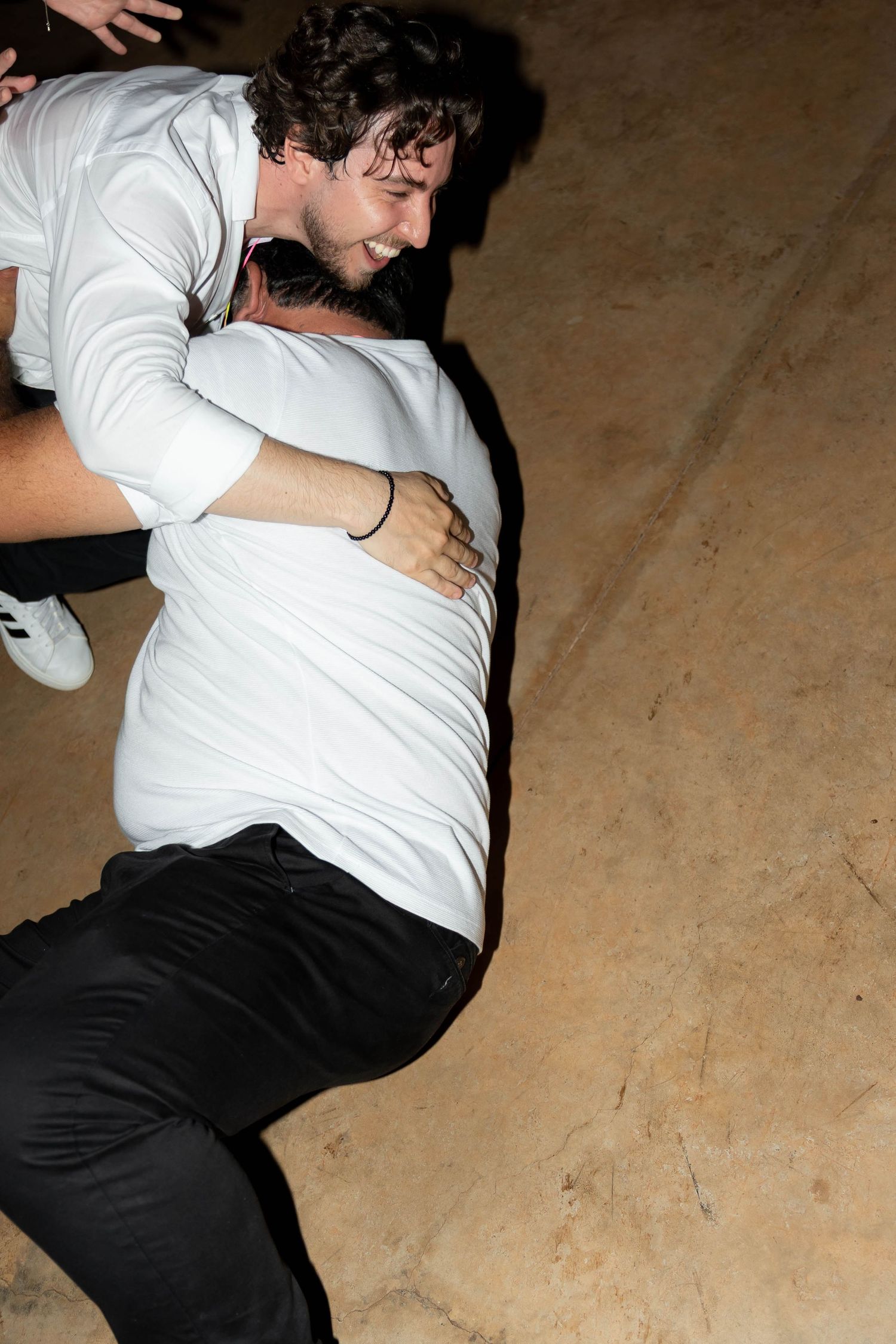 Two young men in white shirts embracing and laughing together at an outdoor evening event.