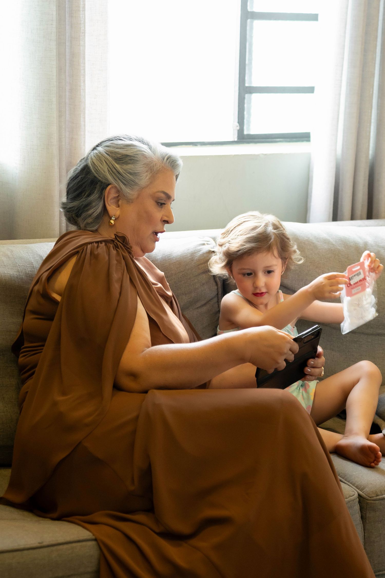 Elderly woman in brown dress sits on sofa with toddler holding a book near a bright window.